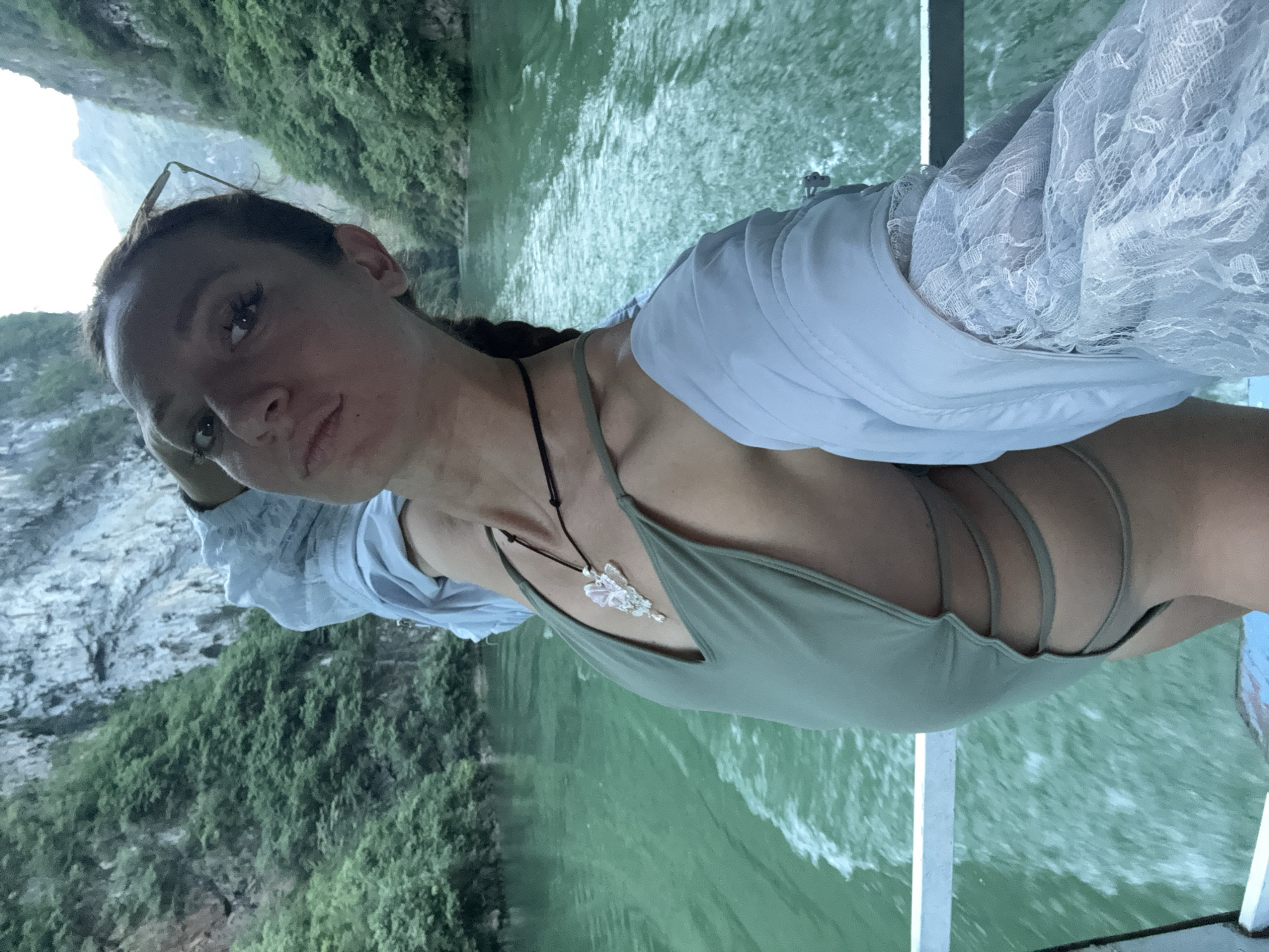 A woman stands on a boat traveling along a body of water with lush, green cliffs rising in the background. She wears a sage green swimsuit with multiple straps and a light blue button-down shirt. A necklace with a light-colored pendant hangs around her neck. The background suggests a location near Đồng Văn Commune, Vietnam.