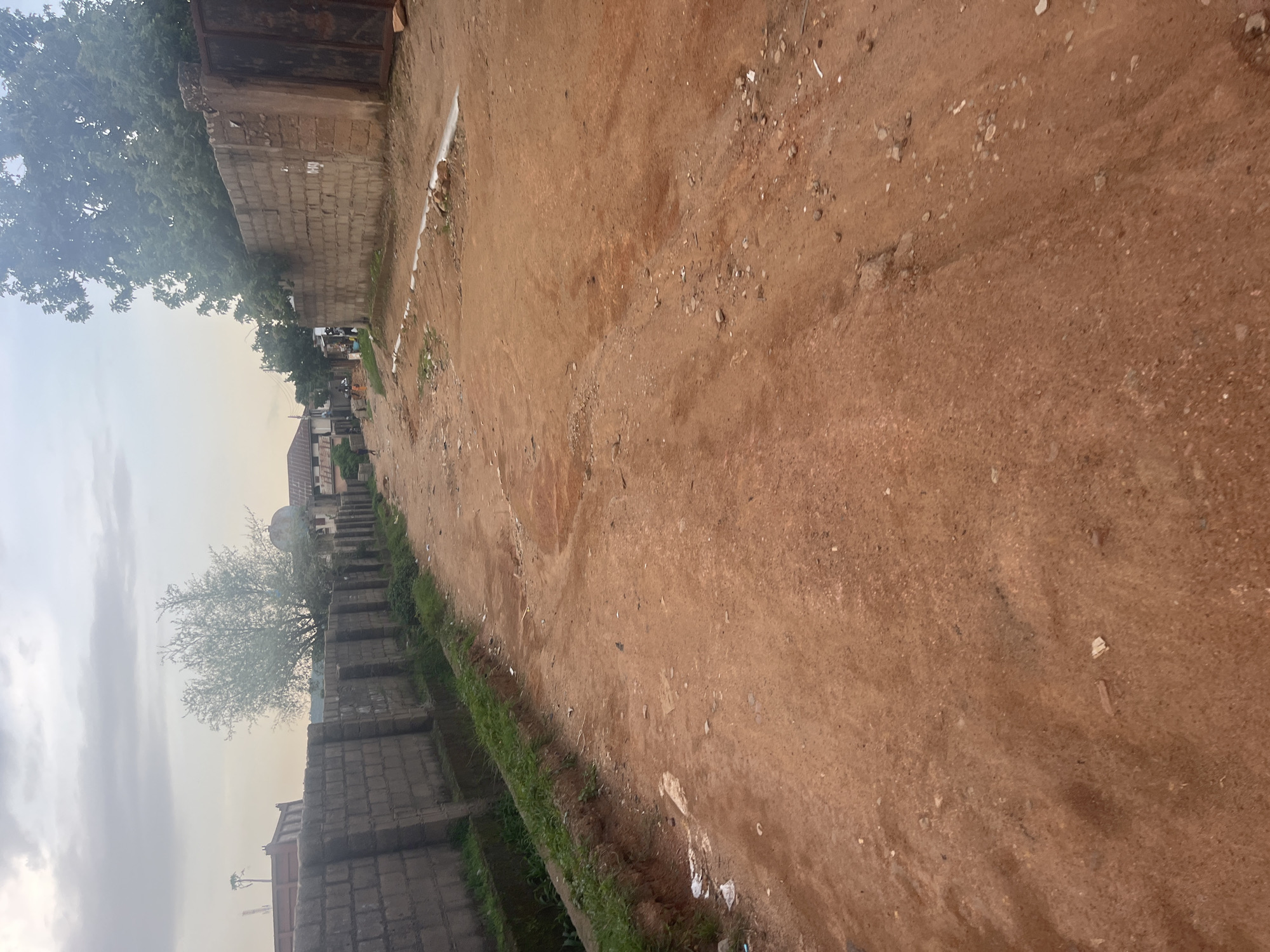 The image depicts a dusty, unpaved road in a rural or semi-urban area. The road surface is composed of reddish-brown soil with scattered small stones and debris. To the left of the road, there is a low wall constructed from concrete blocks, partially covered by grass along its top edge. Beyond the wall, trees with green foliage are visible, and in the distance, there are buildings with pitched roofs, suggesting a residential area. One building in the distance has a satellite dish. The sky is overcast, indicating a cloudy day, possibly near dawn or dusk due to the soft lighting. There are no people or animals actively engaged in activity within the frame, and the scene appears still and quiet. The overall impression is one of a typical, undeveloped street in a Nigerian town, possibly Jos, given the location context. The image is oriented sideways.