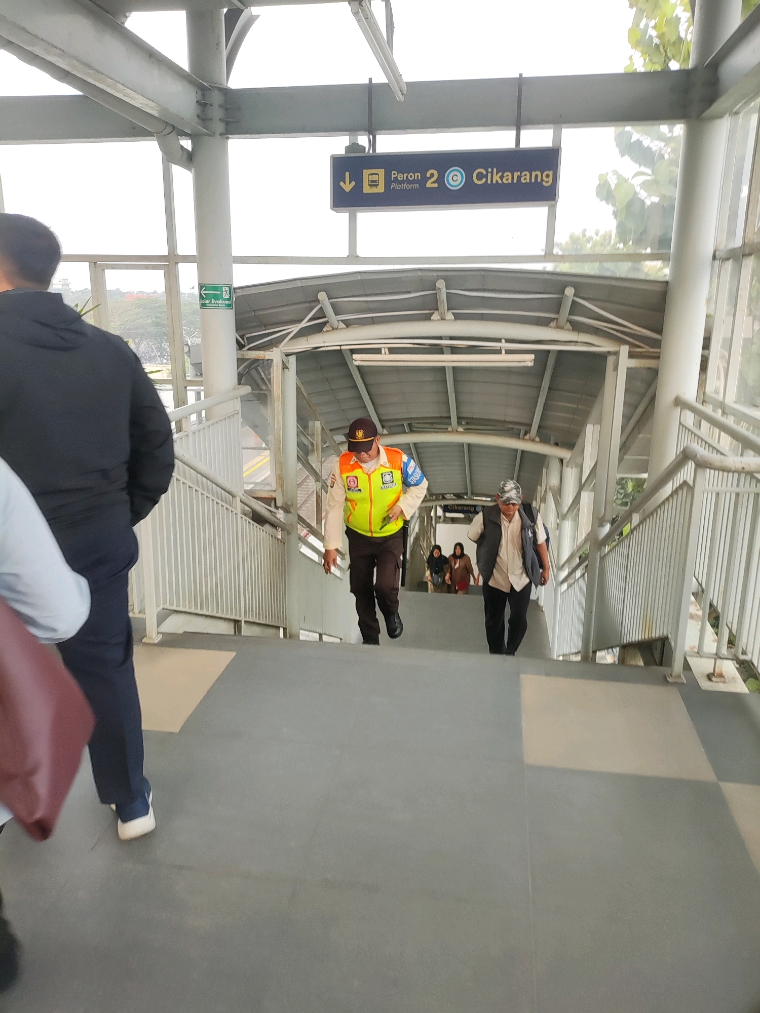 This is a low-angle, eye-level shot of an outdoor pedestrian overpass at the Telagamurni train station in Telagamurni, Indonesia. The overpass is constructed from steel beams and concrete, with white metal railings lining the stairs. A sign at the top indicates "Peron 2 Cikarang" (Platform 2 Cikarang). Two men in uniform are descending the stairs. The man on the left wears a high-visibility vest over a light-colored shirt and dark pants, and a brown baseball cap. The man on the right wears a dark vest over a cream-colored shirt and dark pants, and a patterned baseball cap. Several other individuals, including two women in headscarves, are further down the stairs. A green emergency exit sign is visible on a white pillar to the left. The background shows trees and a bright sky. The stairs and walkway are made of a light gray material, with some sections appearing to be a lighter beige. The image is captured during daylight, with shadows indicating the sun is present.