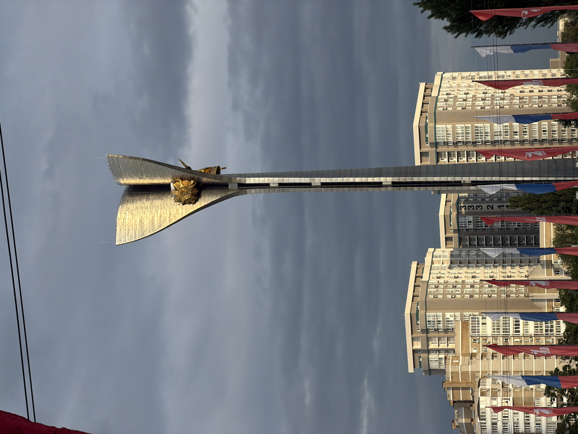 The image captures a vertical view of the Liberation of Rostov-on-Don Monument in Rostov-on-Don, Russia. The monument, featuring a large silver metallic structure with a golden statue at its base, dominates the foreground. Buildings with multiple stories stand in the background, bordered by a display of red, white, and blue flags. The sky is overcast with dark gray clouds. Utility lines are visible on the left.
