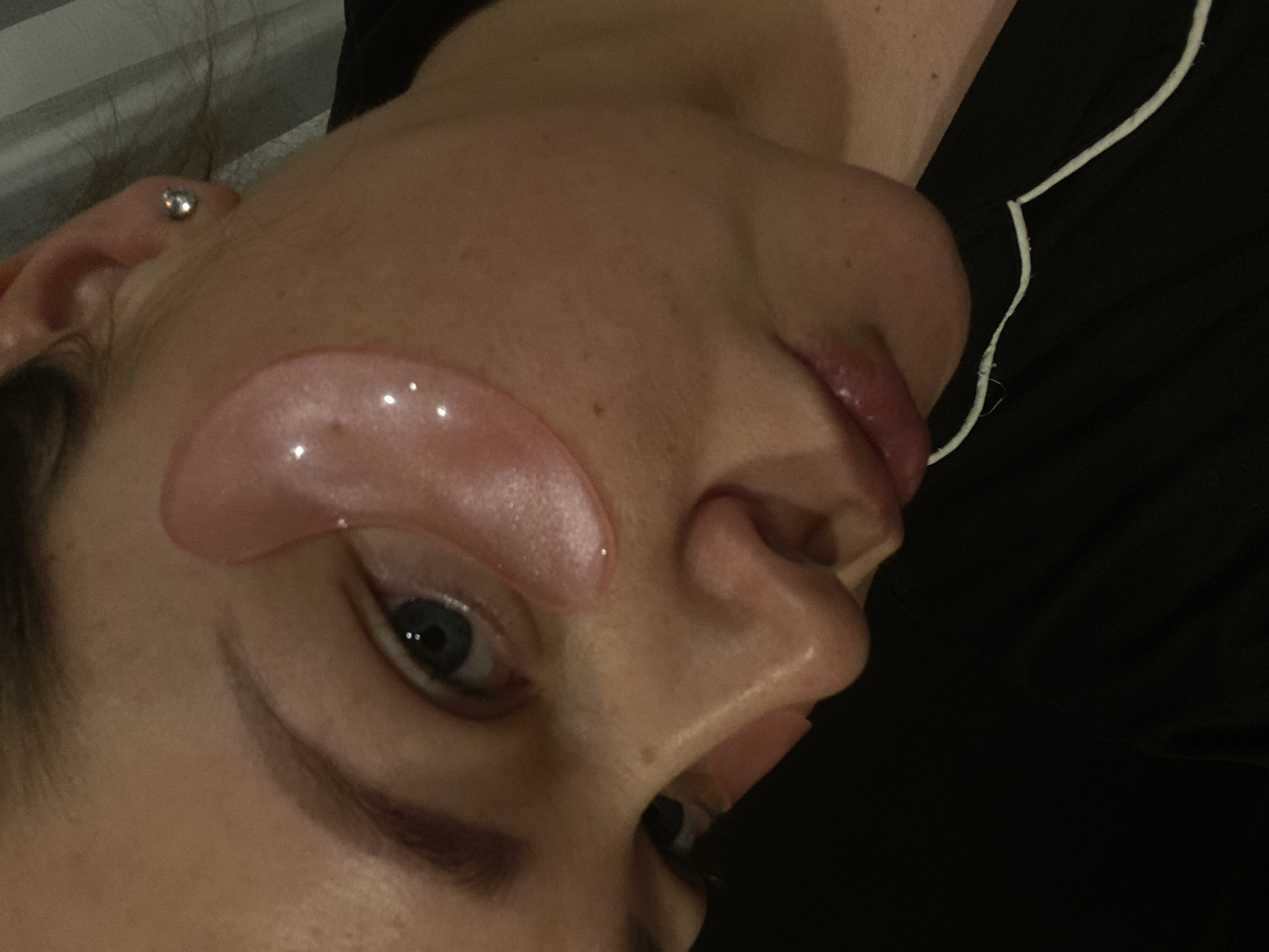 A close-up image features the right side of an adult female's face, angled slightly downwards and to the left. Her right eye, with a visible blue iris, is partially open and looks towards the viewer. A translucent, glossy, pink hydrogel eye patch is affixed beneath her right eye, extending towards the temple, reflecting light. A second similar patch is partially visible under her left eye. Her fair skin shows minor imperfections. A silver stud earring is present in her right earlobe. Her lips are slightly parted, revealing natural lip color. She is wearing a dark-colored garment, likely sleepwear, with a thin white cord or trim visible along the fabric edge near her chin. The background is dark and indistinct, with a subtle vertical striped pattern visible in the upper right quadrant, indicating an indoor environment with soft, directed lighting. The scene is located in the city of Sochi, Russia.