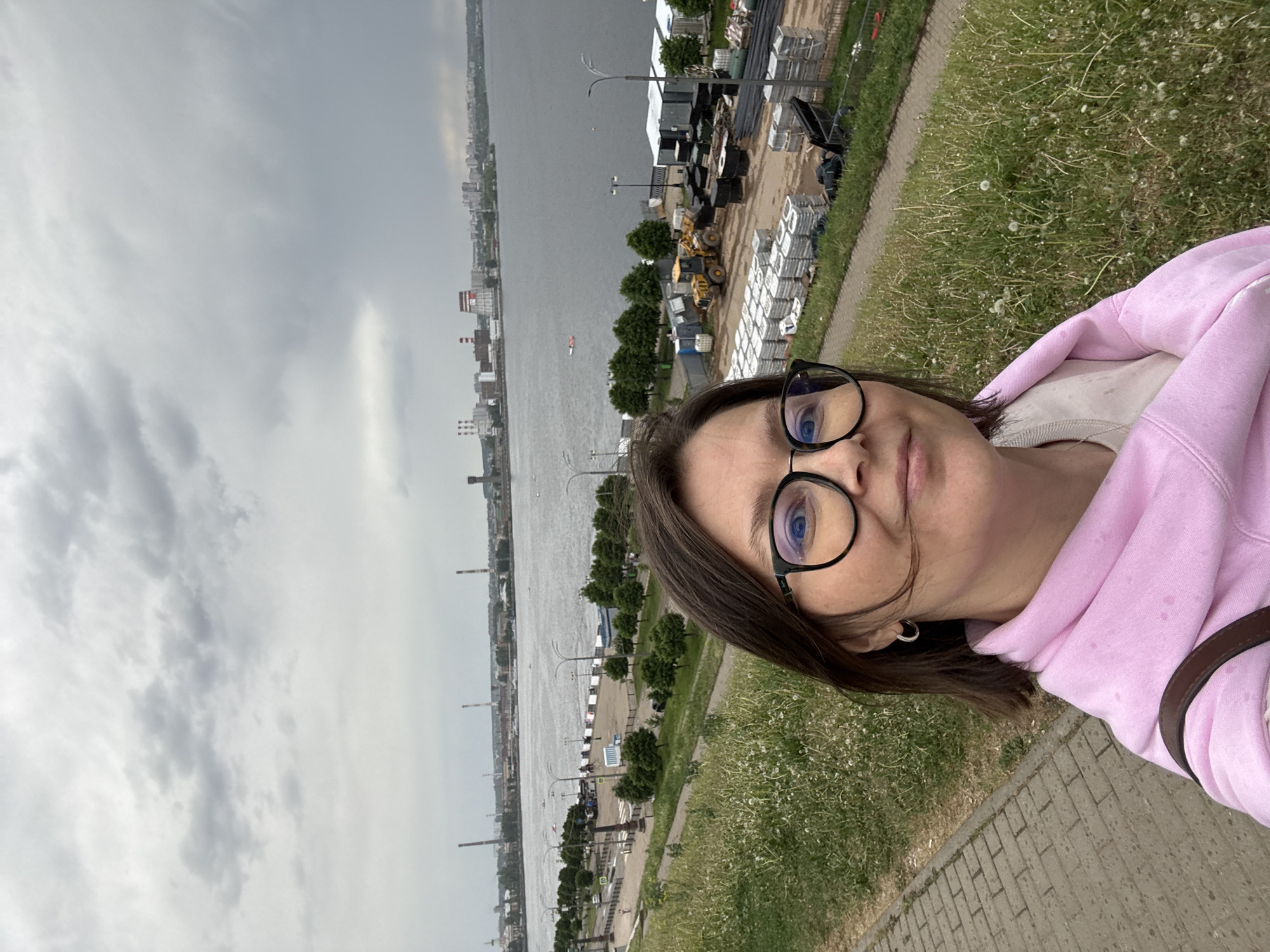 The image is a selfie taken by a woman with glasses and a pink hoodie, likely on a cloudy day. The background reveals a cityscape along a river, with industrial elements such as chimneys and cranes. There's also construction equipment and materials visible in the background. The photo was taken along the Central Embankment near the Izh River in Izhevsk, Russia. The scene shows a paved walkway bordering a grassy area with scattered dandelions.