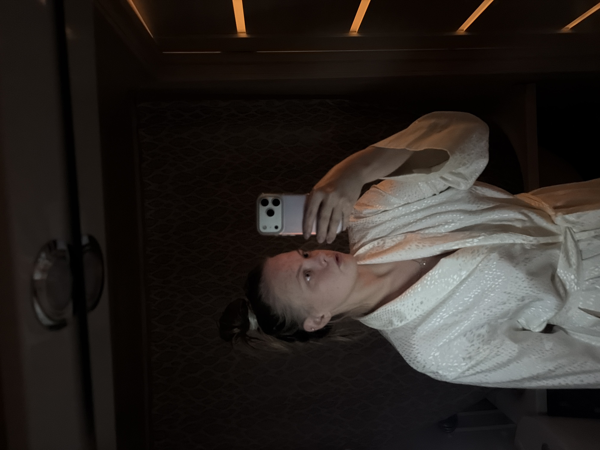 A woman is captured in a selfie, taken from her upper torso upwards, in a dimly lit indoor setting. She is wearing a white, textured robe, possibly made of terry cloth or a similar material, with a subtle patterned design across it. Her dark hair is pulled back, revealing a small stud earring in her left ear. She holds a light-colored smartphone with a distinct camera array in her right hand, positioned to take the photograph. Her gaze is directed towards the phone's camera, with a neutral expression.
The background is dark and appears to be an interior space, possibly a hotel room, spa, or dressing area, in Izhevsk, Russia. To the left of the frame, a light-colored wall or door is visible, featuring a circular, metallic handle. The main background wall has a dark, textured pattern, possibly wallpaper or a decorative panel. Above, narrow, rectangular light strips illuminate the wooden ceiling, casting a warm glow. The overall lighting is low, creating a somewhat intimate or relaxed ambiance. No visible text is discernible in the image.