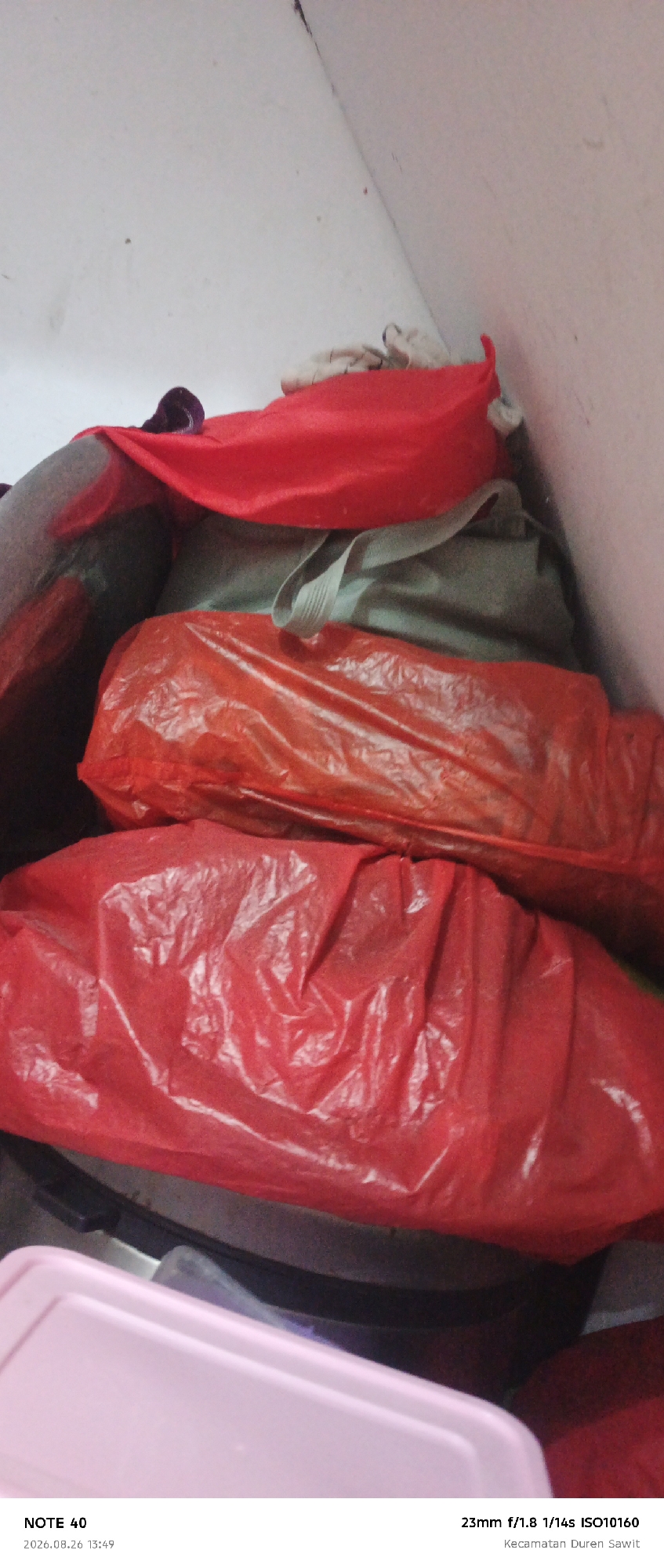 The image shows a stack of bundled items wrapped in red and orange plastic. The items are piled on top of each other in a cramped space with white walls. A portion of a pink plastic container is visible at the bottom of the frame. The timestamp "2026.08.26 13:49" and location "Kecamatan Duren Sawit" suggest the image was taken in Duren Sawit District, East Jakarta, Indonesia.