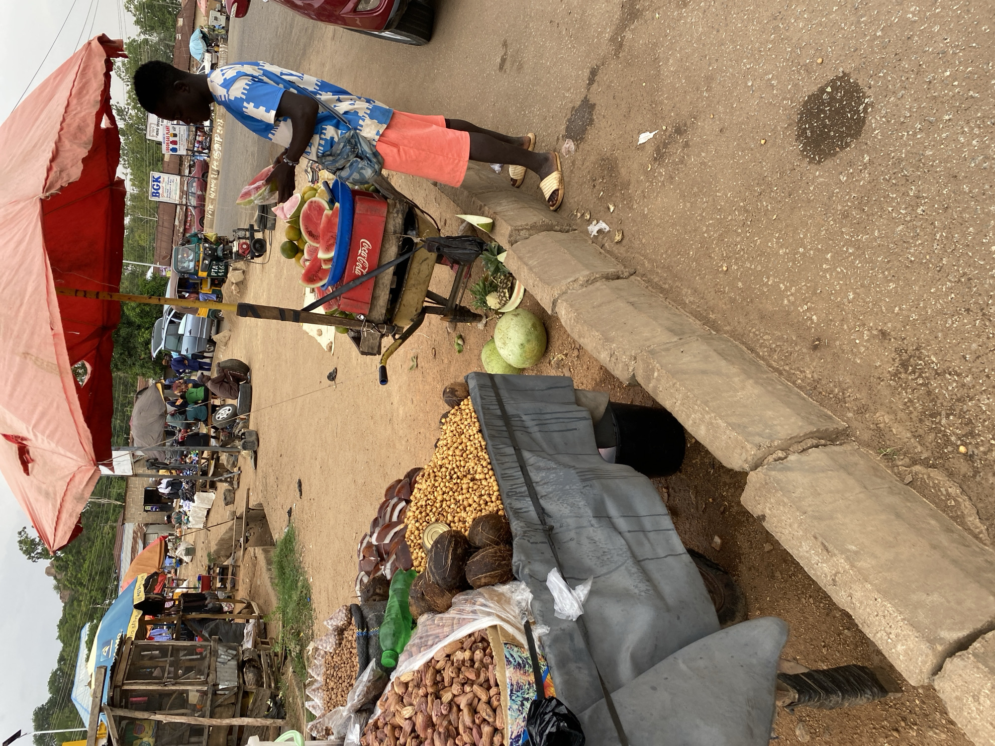 A young man in a blue patterned shirt and orange shorts stands beside a cart filled with watermelon slices inside a red Coca-Cola crate. He appears to be preparing to serve a customer, likely at a roadside stall in Bukuru, Nigeria. A large red umbrella offers shade. The cart is parked on a worn curb next to a collection of nuts and coconuts covered by a grey tarp. In the background, there are other roadside stalls, vehicles including a "keke napep" tricycle taxi, and various signs (BGK, LIBGON GAS PLANT, etc.).