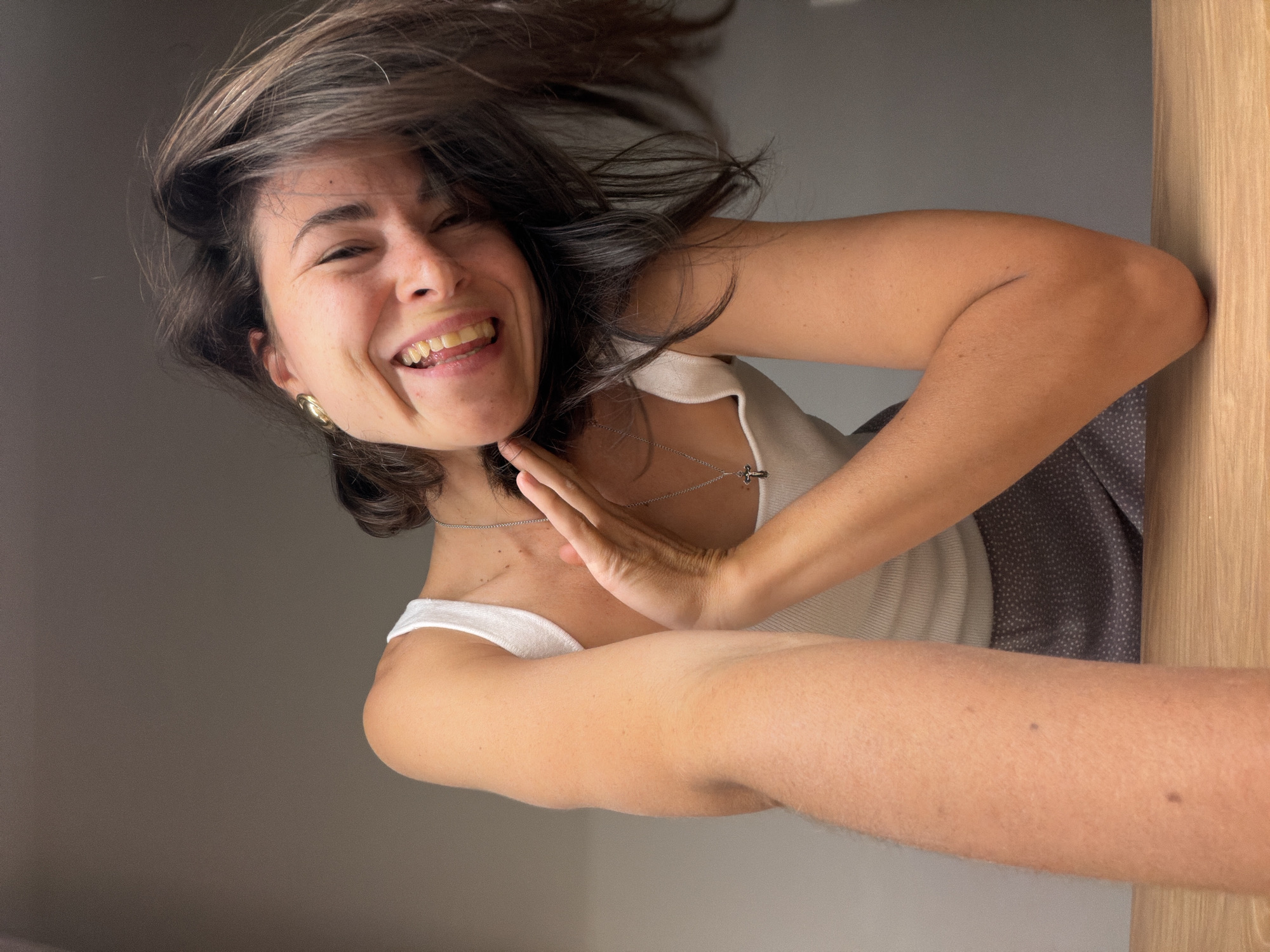 A woman with dark hair and a white tank top is smiling and looking down at the camera. She has fair skin and is wearing a necklace and earrings. Her hair is slightly disheveled, suggesting movement. Her hand is raised near her chin. The background is a mix of gray and wooden surfaces. The location in the city of Budva, Montenegro, cannot be determined from the image.