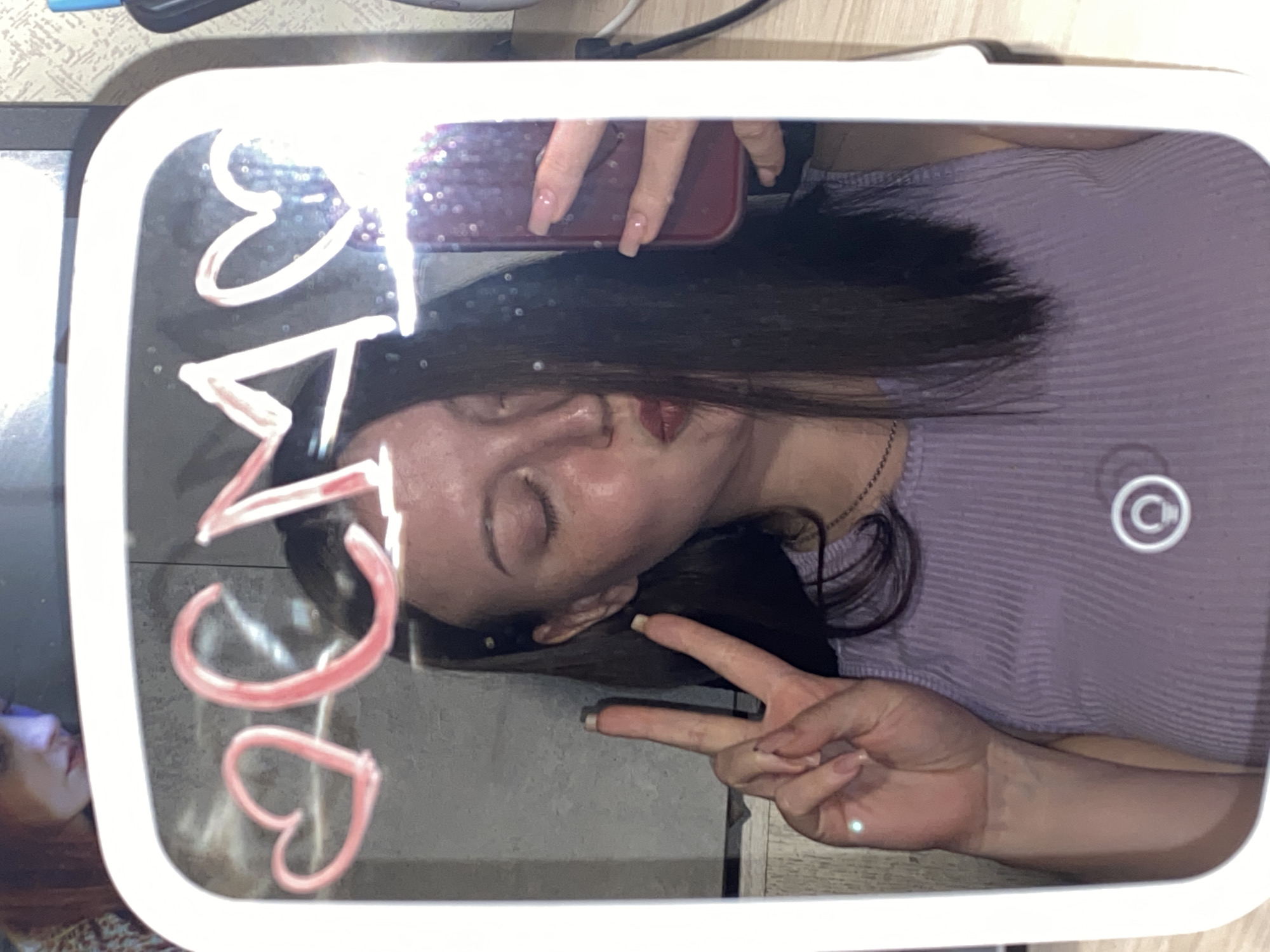 The image shows a reflection in a lighted mirror with a woman posing. The woman has long, dark hair and is wearing a light purple striped shirt. She's holding up a peace sign with her right hand. She is also holding a phone in the upper part of the mirror. On the mirror, the word "CAT" is written vertically, accompanied by heart shapes. A partial image of a person is visible to the left of the mirror, likely on a surface where the mirror is placed. The environment suggests an indoor setting, possibly a bedroom or dressing area. There's no information to determine the location in the city of Chaikovsky, Russia, or whether the image was even taken there.
