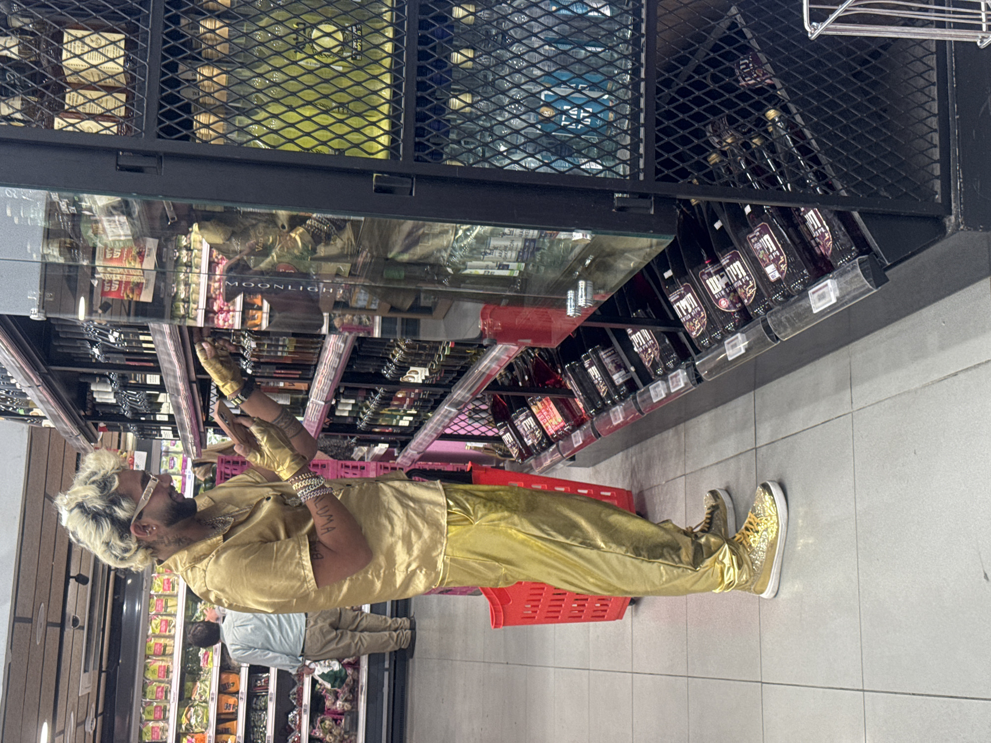 A person dressed in a shiny gold shirt, pants, and shoes stands next to a red shopping basket while browsing the shelves in a supermarket. The supermarket shelves display various food items, including wine and other packaged goods. The location is likely a supermarket in Givatayim, Israel, although specific indicators in the image are limited. Another person in a light blue shirt and khaki pants is visible in the aisle behind the main subject.