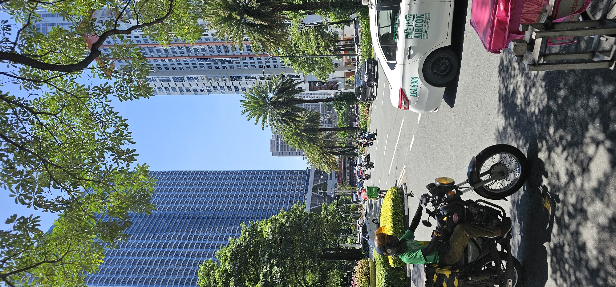 This image captures a street scene in Makati, Philippines, under a clear blue sky. In the foreground, a person wearing a green shirt and helmet rides a black motorcycle with yellow and black cargo boxes. The motorcycle has a visible license plate and tire. To the right of the motorcycle, a white car with the license plate "AGA 8303" is parked. The car has text on its side, including "AIRCON" and a phone number. Further down the road, other vehicles, including motorcycles and cars, are present.
The background features tall buildings, characteristic of an urban environment. The building on the left is a tall, blue skyscraper with many windows. The building on the right is also a high-rise, with a lighter facade and visible windows. Palm trees and lush green trees line the street, providing shade and a tropical feel. The road is paved asphalt, and the scene is illuminated by bright sunlight, casting shadows on the road from the trees. The overall impression is a busy, sunny day in a modern city.