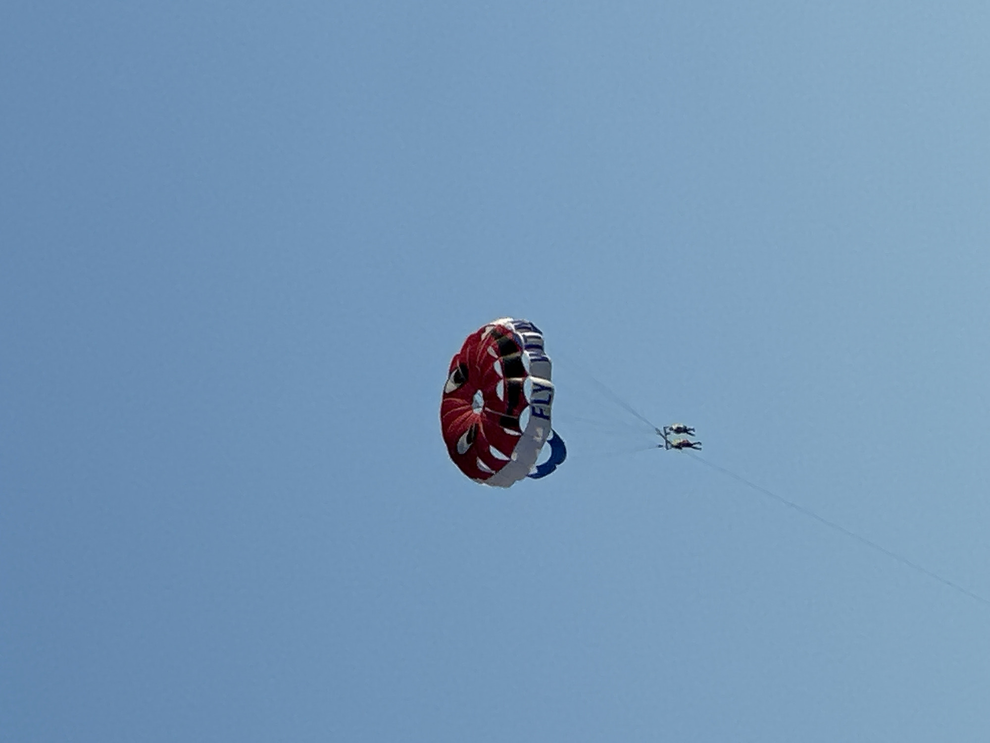 A parasail with a red and white canopy, featuring the word "FLY" in blue lettering, is suspended in a clear blue sky above Budva, Montenegro. Two figures are visible, suspended below the parasail by a tow rope, suggesting they are engaged in parasailing. The vast expanse of the sky dominates the image, with no other discernible features of the city or landscape visible.
