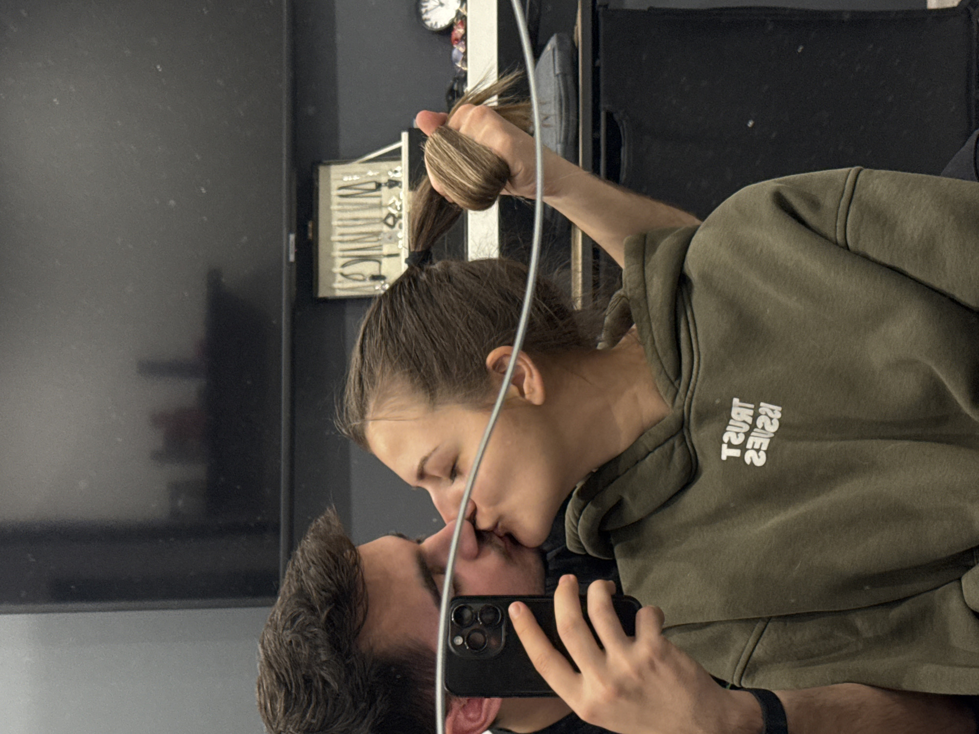 A reflection shows a man and a woman kissing. The woman is wearing an olive green hoodie with the words "TRUST ISSUES" printed in reverse. She has brown hair pulled back in a ponytail, and she is holding the end of her ponytail in her hand. The man has short, dark hair and is holding a black smartphone, presumably taking the picture in the reflection. The background includes a dark gray wall, a gray surface, a jewelry box with hanging necklaces, a clock, and a dark appliance. There is no indication of the city of Sofia, Bulgaria.