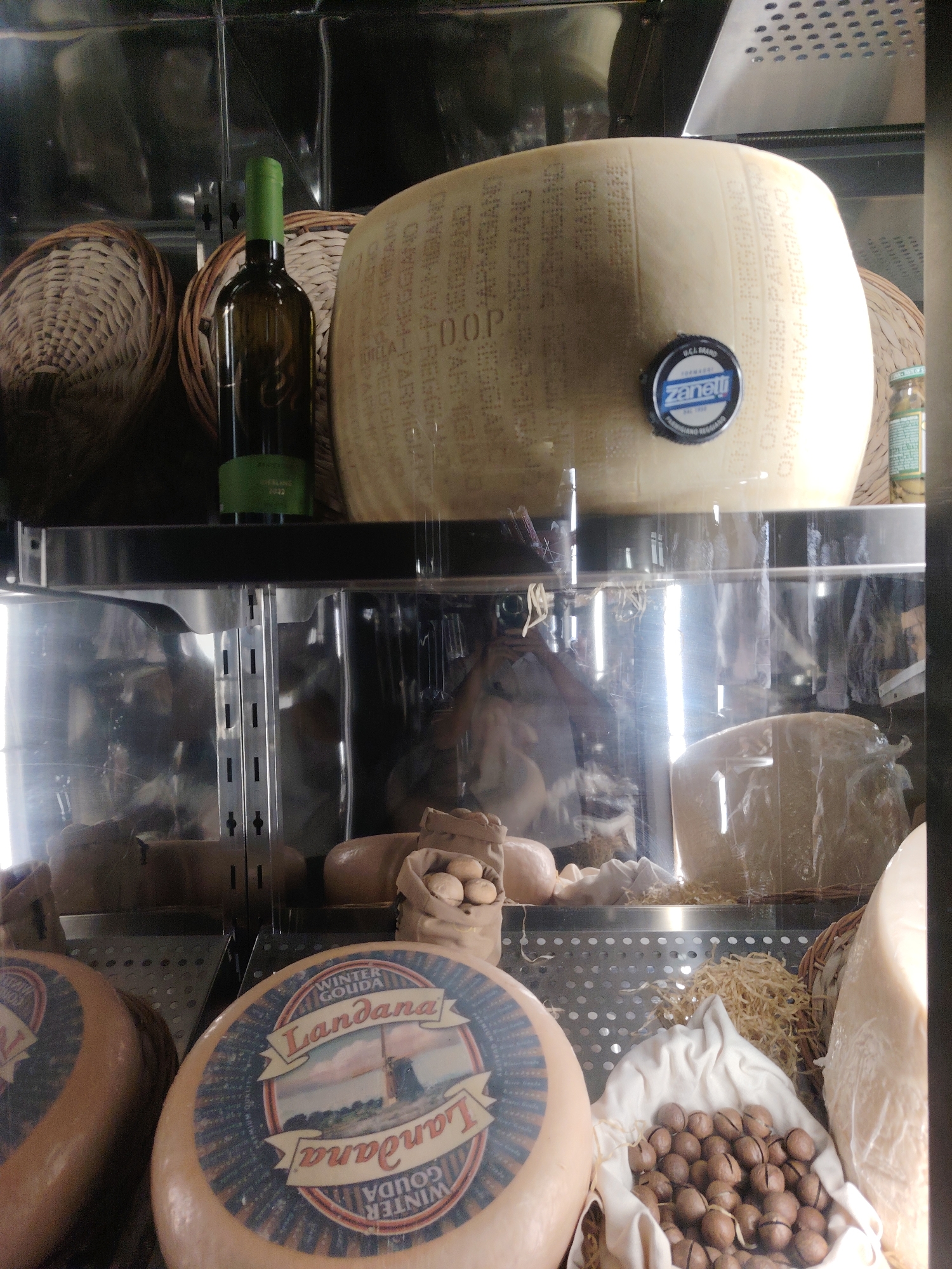 The image depicts a display of various cheeses and other food items within a refrigerated case. In the upper section, a large wheel of Parmigiano-Reggiano cheese is prominently displayed, featuring a "Zanetti" brand stamp. To its left, a green-bottled wine is positioned next to two woven baskets. Below, a variety of cheese wheels are arranged, including two labeled "Landana Winter Gouda." Also visible are bags of nuts, possibly macadamia nuts, and portions of other cheeses. The setting appears to be a shop or market, potentially in a city like Konigil, Uzbekistan, given the context provided. The lighting is artificial, and reflections on the glass casing are noticeable, showing hints of the surrounding environment.