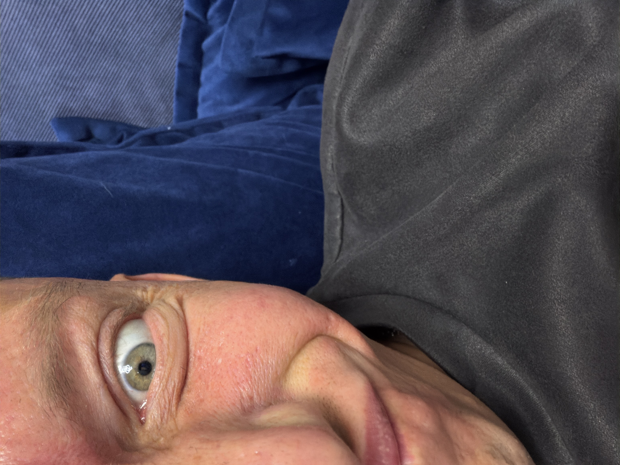The image is a close-up, low-angle selfie taken indoors. The subject is a person with fair skin and light-colored eyes, looking directly at the camera with a surprised or playful expression. Their left eye is prominently featured, with a dilated pupil. The person is wearing a dark gray crew-neck t-shirt. In the background, a plush blue fabric, possibly a couch or blanket, is visible. The lighting is somewhat harsh, creating highlights on the skin. There is no discernible information about the location within Moscow, Russia, or any other specific details of the environment.