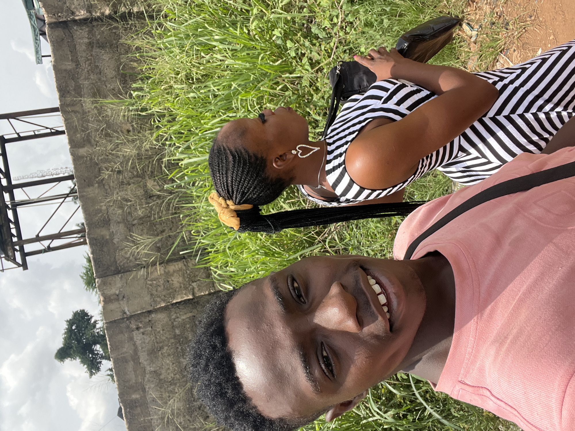 Two individuals are in the city of Obosi, Nigeria. A male, wearing a pink shirt, faces the camera, smiling. Behind him, a female is lying on her back, wearing a black and white striped dress. She is looking upwards. She has braided hair tied back with a gold scrunchie, and is wearing heart-shaped earrings. She is holding a black bag in her arms. They are in a grassy area near a concrete wall. Metal scaffolding is visible in the background along with trees and sky.