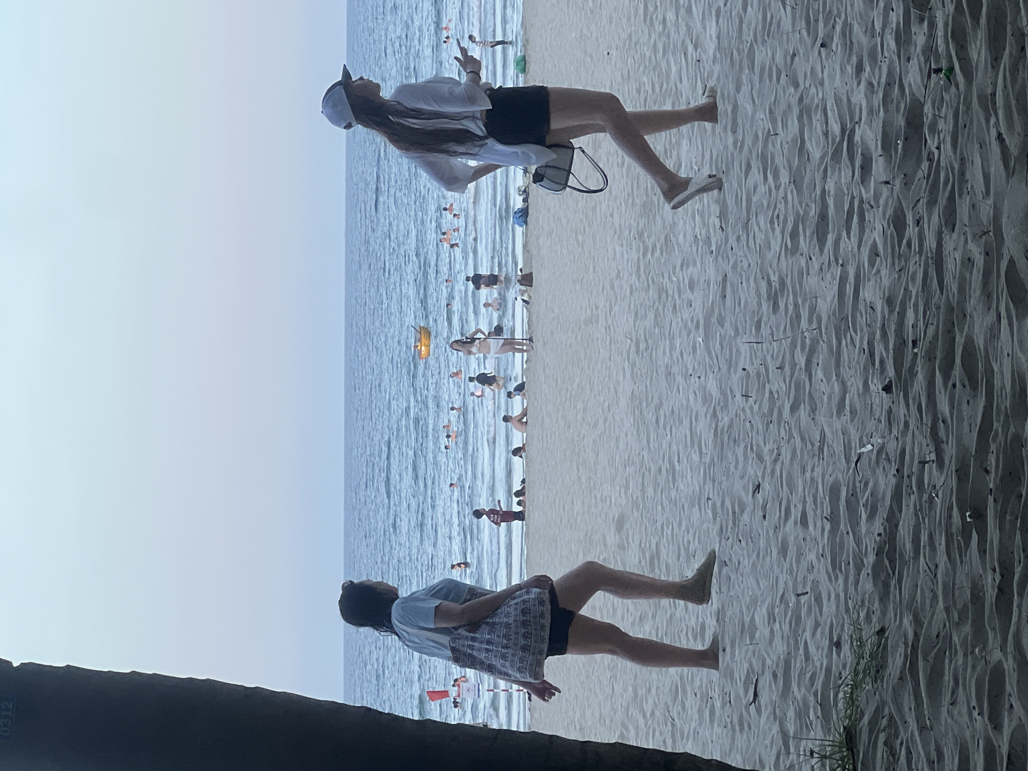 The image shows a beach scene at dusk in Da Nang, Vietnam. Two women are walking on the sand, with the ocean and other beachgoers in the background. The woman on the left is wearing a light blue t-shirt, dark shorts, and carrying a patterned tote bag. The woman on the right is wearing a white, long-sleeved shirt, black shorts, and a white baseball cap. The ocean is calm with several people swimming and playing in the water. A yellow boat is visible in the distance. The sand is light brown and appears to be rippled. A dark tree trunk is partially visible on the left side of the image. The sky is a pale blue, indicating twilight.