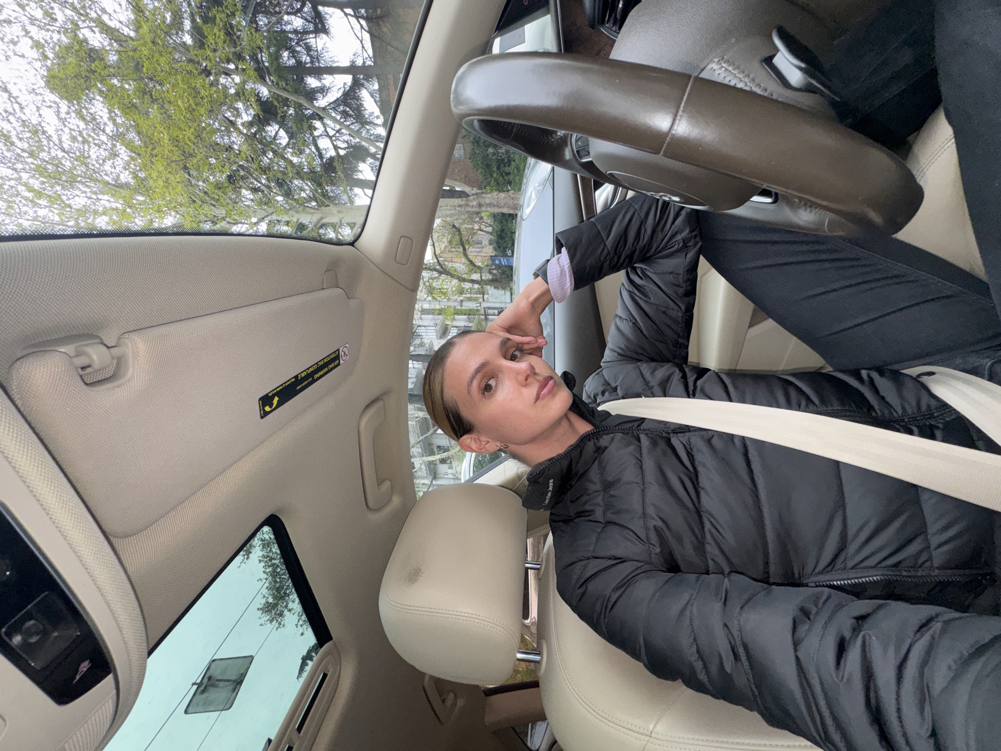 A selfie photograph, rotated 90 degrees counter-clockwise, captures a young woman seated in the driver's seat of a car. She looks directly at the camera with a neutral expression, her light brown hair pulled back. She is wearing a black quilted jacket, with "Calvin Klein" faintly visible on it, and a lighter long-sleeved shirt underneath. A light-colored seatbelt crosses her chest, and her right hand rests on her temple.
The car's interior features beige leather seats and headrests, one of which has a dark smudge. A dark brown steering wheel, a light beige headliner, and a sun visor are also visible. An overhead console with controls is positioned above the windshield.
Through the car's windshield and open sunroof, an exterior scene is visible. Trees with fresh, light green leaves indicate a spring setting under an overcast sky. A residential building and parts of another car can be seen in the background.
Visible text on the sun visor includes a warning label reading "ATTENTION: SAC GONFLABLE" and "AIR BAG INFLATABLE" accompanied by a warning symbol. This scene is set in Tbilisi, Georgia.