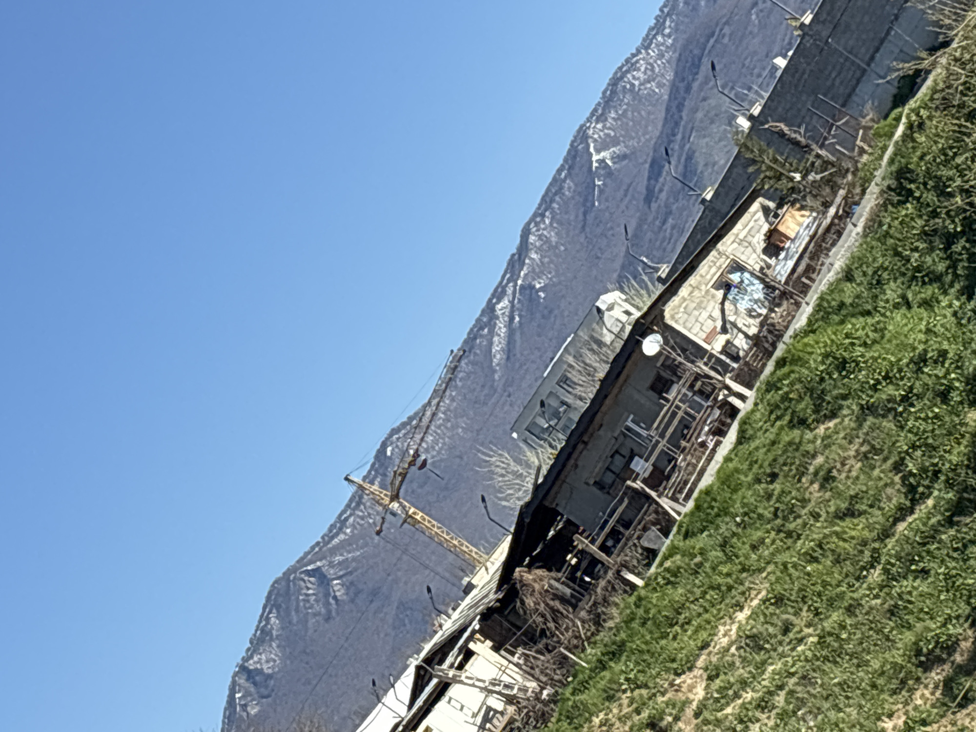 The image shows a construction site in Tbilisi, Georgia, near a mountain. A yellow crane is visible next to the unfinished building, which is surrounded by scaffolding. The foreground is grassy, and a fence runs along the edge of the construction site. A satellite dish is affixed to the building. The sky is clear and blue.