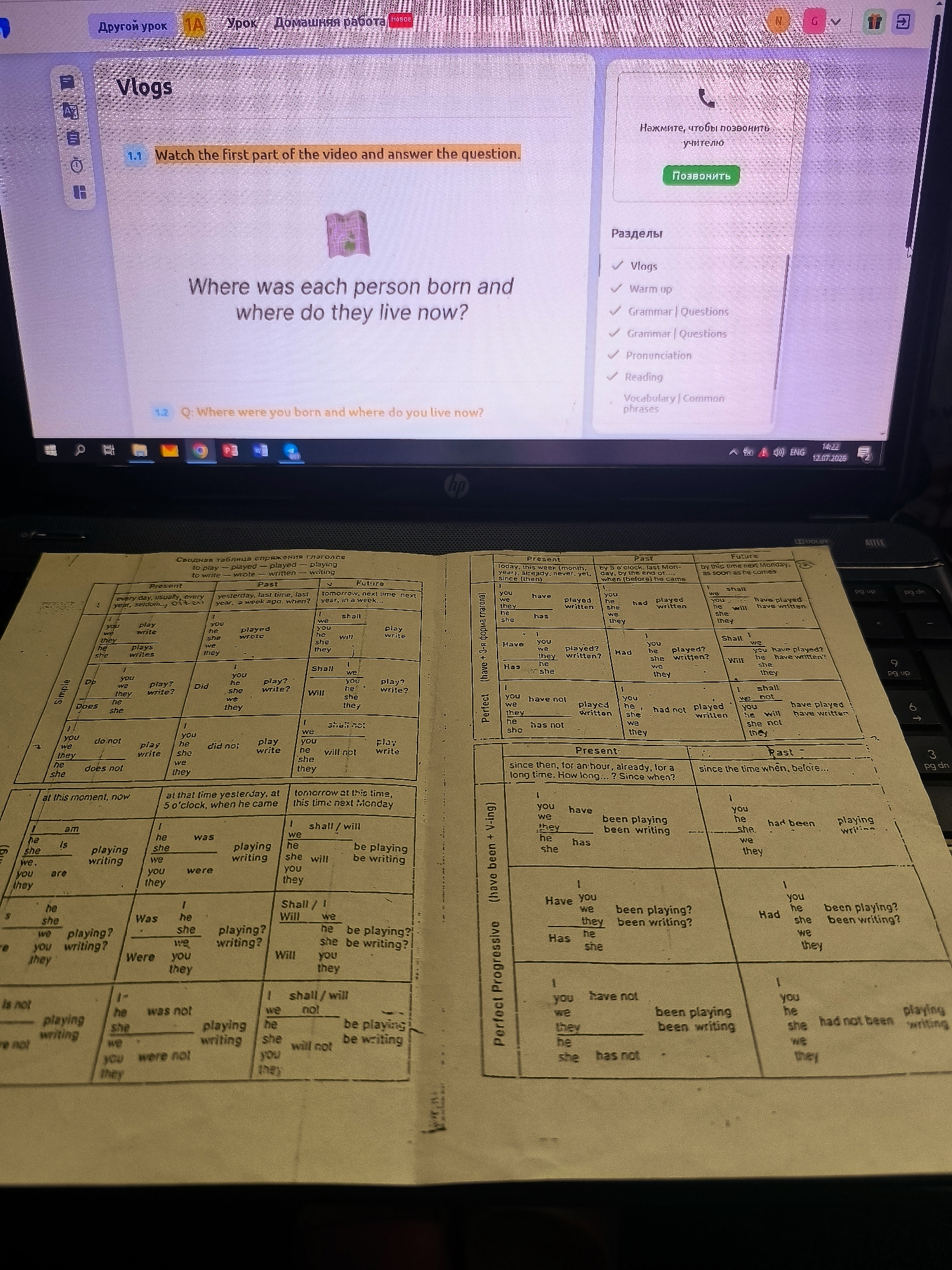 The image shows a laptop screen displaying a webpage with the title "Vlogs" and an interactive lesson. To the right of the screen, a portion of a handwritten English grammar chart is visible, detailing verb conjugations across different tenses (Present, Past, Future) and aspects (Simple, Perfect, Perfect Progressive). The chart contains examples of regular and irregular verb forms. The laptop is positioned on a table. The background is blurred, but it suggests an indoor setting. There is no information in the image that allows for the identification of the city of Almere, Netherlands, or any specific location within it.