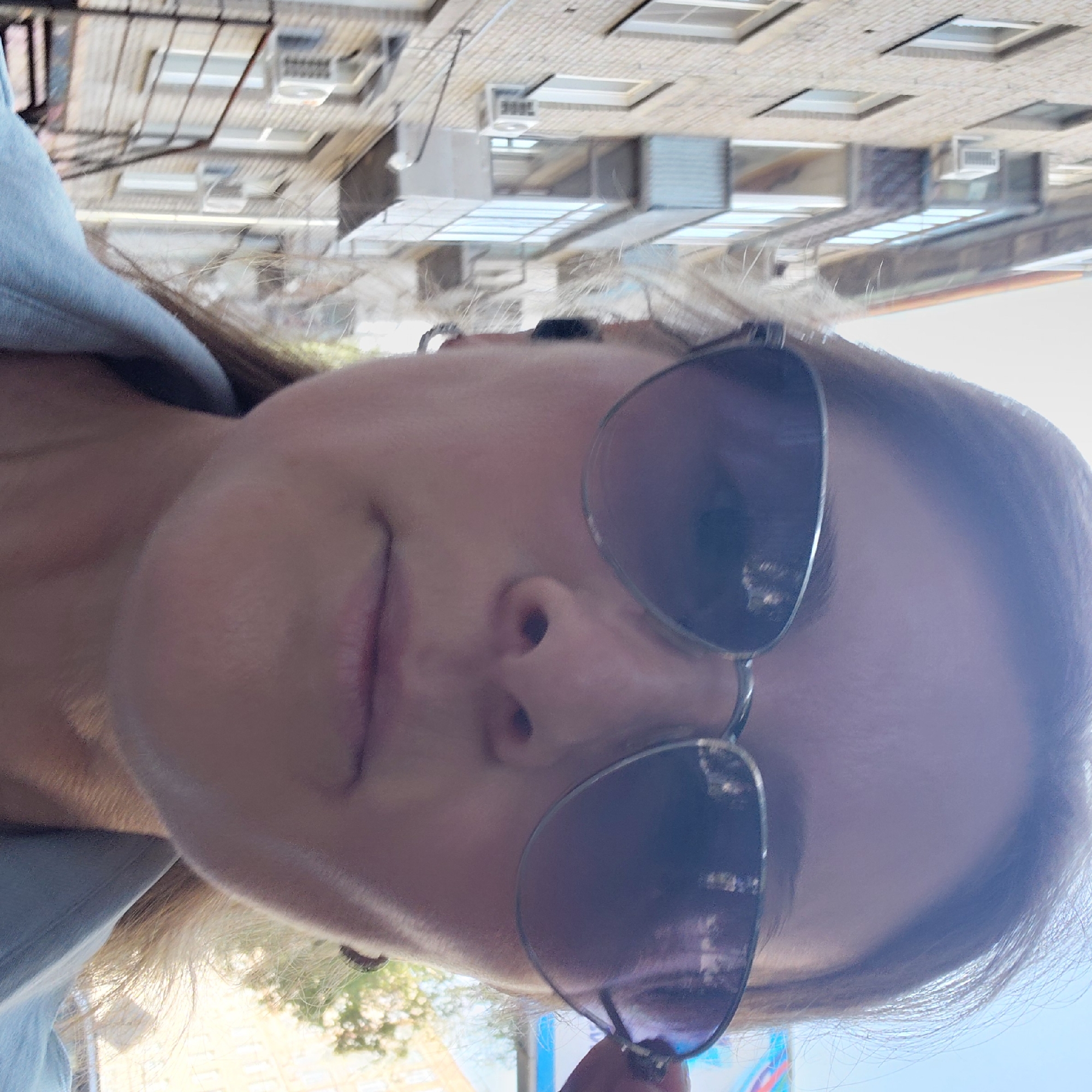 The image is a close-up selfie of a young woman wearing sunglasses and a light blue collared shirt. She has blonde hair and is wearing a small hoop earring in her left ear. The background shows the facade of a brick apartment building with several windows and balconies, and an air conditioning unit is visible on the building. There are also trees and sky visible in the background. The scene appears to be an urban environment, consistent with a street in Novosibirsk, Russia.