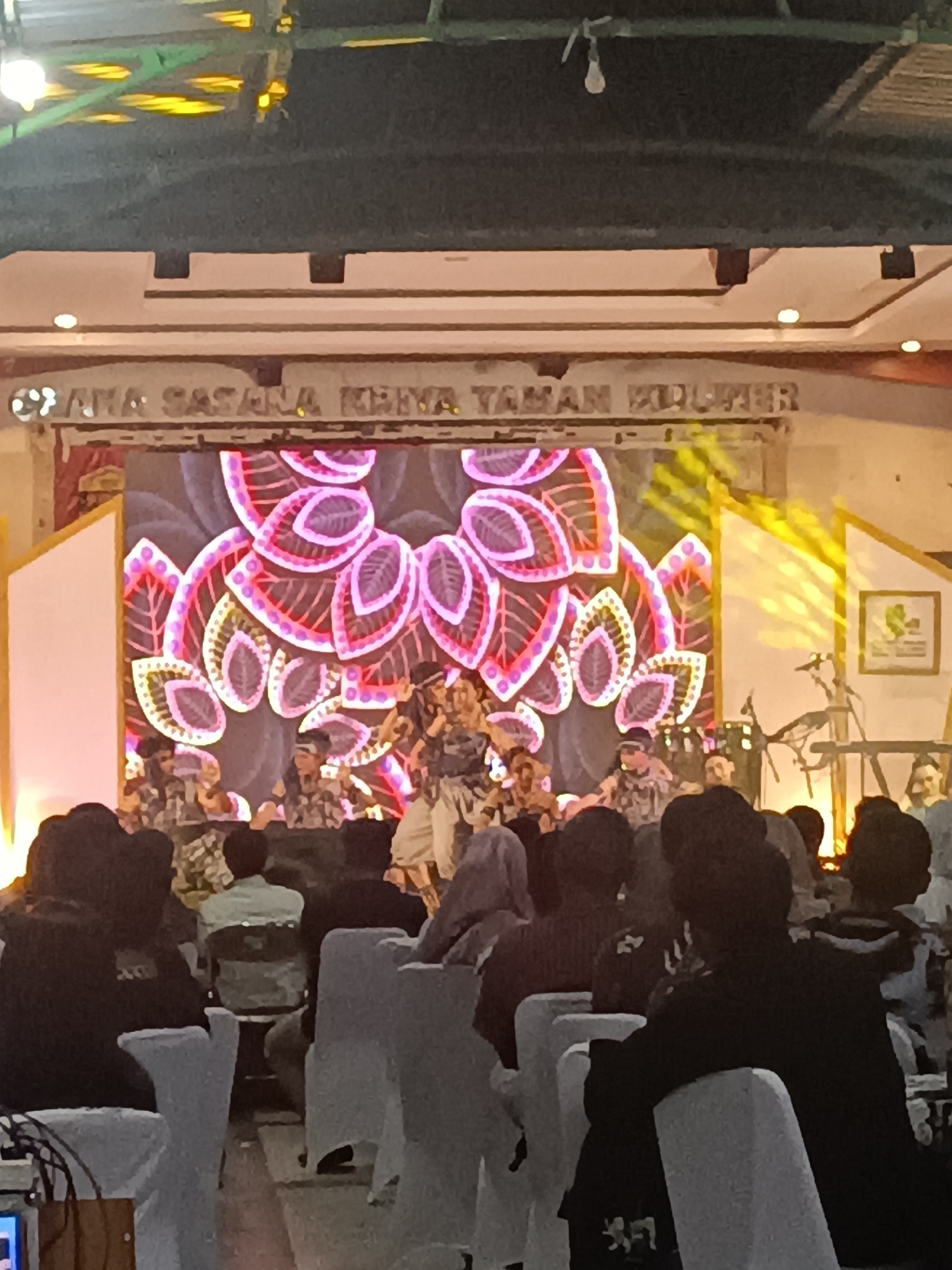 The image depicts an indoor event, likely a performance or ceremony, taking place at the Graha Sasana Krida Taman Kuliner in Condongcatur, Indonesia. In the background, a large LED screen displays a vibrant, abstract pattern resembling stylized leaves in shades of pink and purple, illuminated by stage lighting. Several performers, dressed in traditional Indonesian attire, are on stage, some appearing to be singing or dancing. In the foreground, an audience is seated in rows of white chairs, facing the stage. The overall ambiance suggests a cultural event with a festive atmosphere.