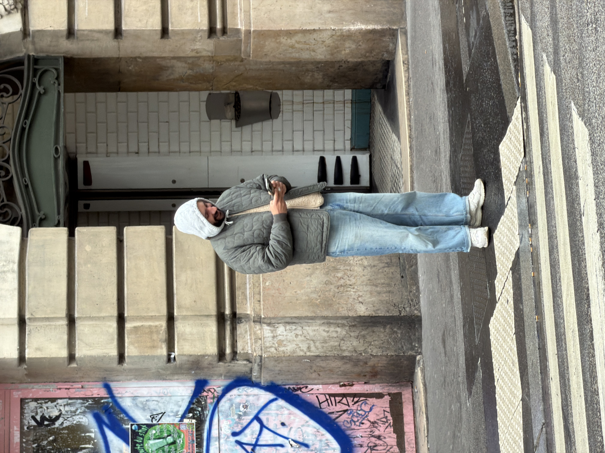 A man in a light grey hooded sweatshirt and jeans stands on a crosswalk next to a building entrance in Paris, France. The building's facade features light-colored vertical architectural details above a wall with graffiti-covered doors. The man's feet are near the edge of the sidewalk, bordering the crosswalk painted in black and white stripes. He wears white sneakers. The building's entrance features ornamental ironwork and light-colored tiled walls.