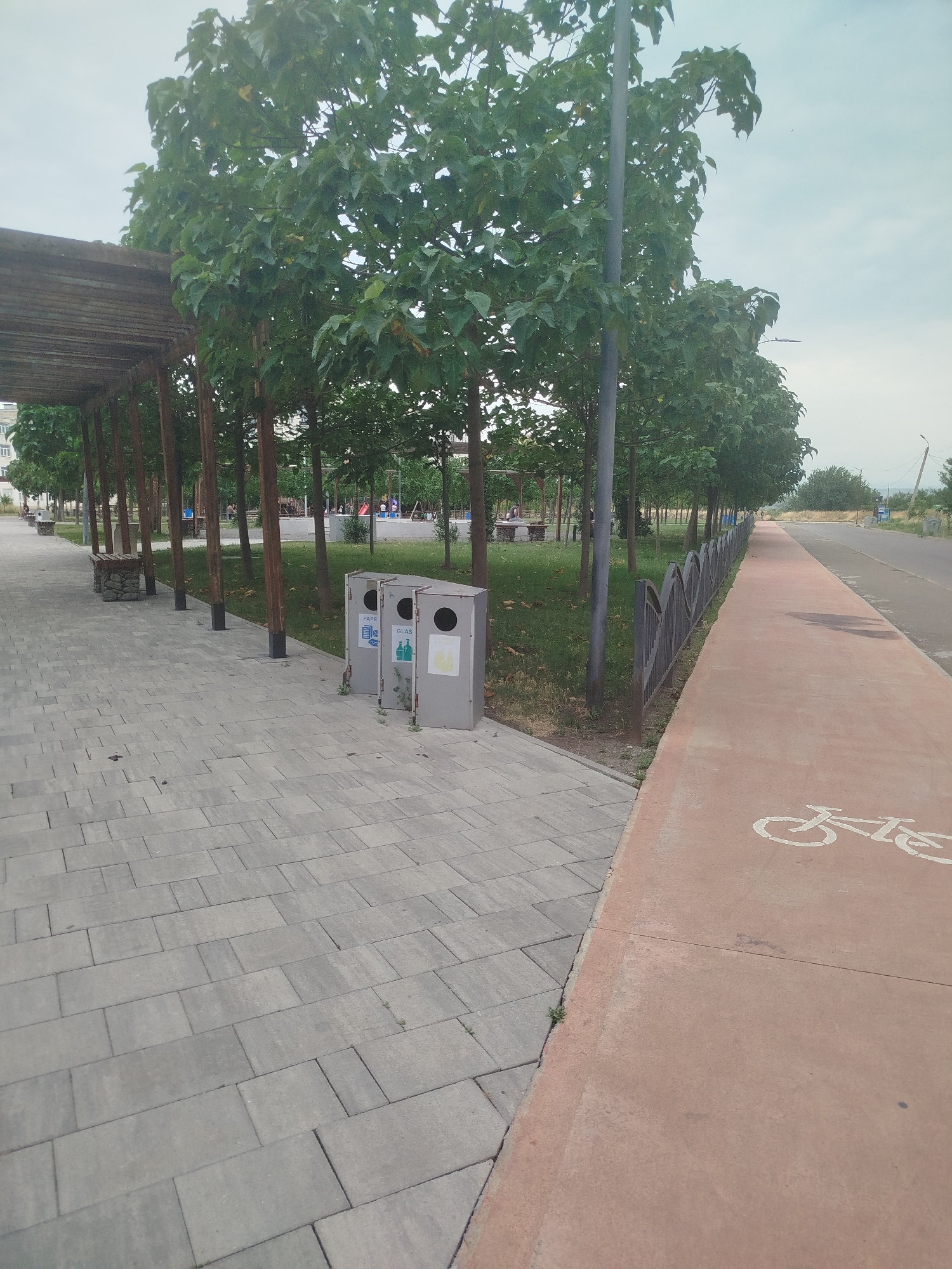 The image captures a scene in Rustavi, Georgia, likely a park or public area. A paved walkway with large, gray rectangular tiles runs along the left side of the frame, leading towards a wooden pergola structure. Beside the walkway are two modern, gray trash receptacles, one labeled "PAPE" and the other "GLAS." Beyond the walkway, a grassy area is dotted with young trees and a few people can be seen in the distance near playground equipment. A dark metal fence with a decorative wavy pattern runs along the edge of the grassy area, separating it from a dedicated bike path. This bike path, painted a reddish-orange, features a white bicycle symbol. The bike path continues into the distance, bordered by a road on the right. The sky is overcast, suggesting a cloudy day.