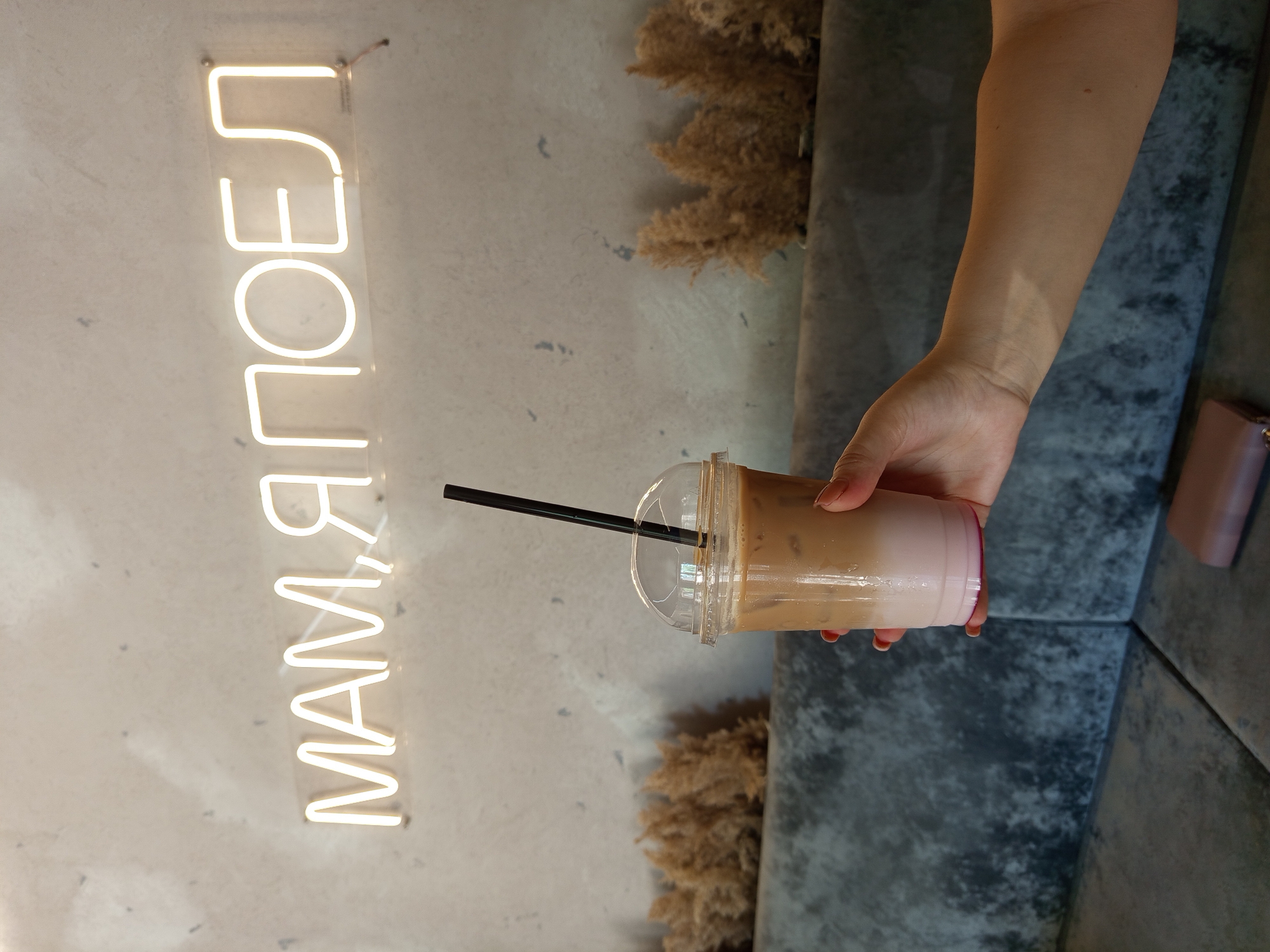 In Bender, Moldova, a person's hand holds a clear plastic cup filled with a layered beverage, likely iced coffee. The drink has a pinkish base, topped with a creamy brown liquid and ice. A black straw is inserted into the cup. The background features a concrete wall with a bright neon sign that reads "МАМ, Я ПОЕЛ" in Cyrillic script, translating to "Mom, I ate." Adjacent to the wall, dry, wispy plant decor is arranged behind a dark grey sofa. A portion of another person's arm and a pink wallet are visible on the sofa in the foreground. The lighting suggests an indoor setting, possibly a cafe.
