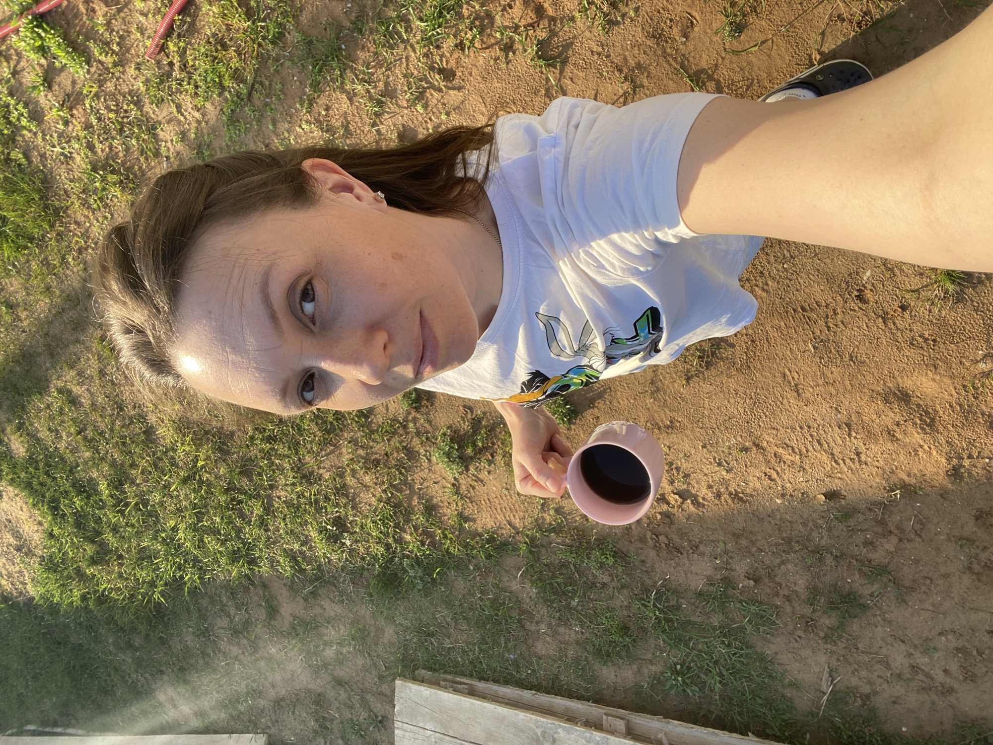 A medium-height fair-skinned woman with light brown hair is holding a pink mug of coffee, likely outdoors in an area near Chaikovsky, Russia. She is wearing a white T-shirt with a cartoon on it. The background includes grass, dirt, and partial view of what might be a wooden structure in the lower left corner of the image. She appears to be in her early to mid-30s. The ground is uneven and sparsely covered with grass. The photo appears to have been taken in bright sunlight, casting shadows throughout the scene.