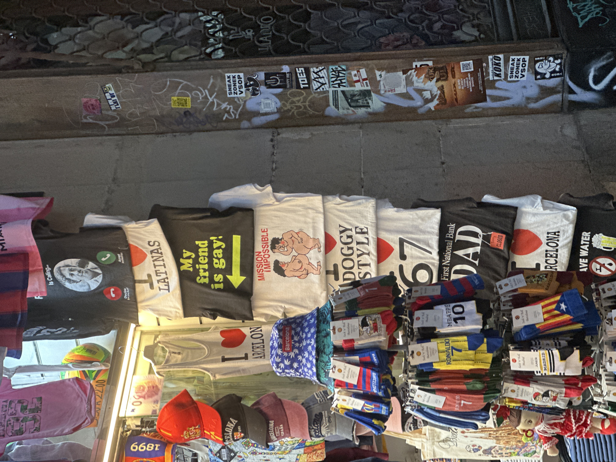 The image depicts a street stall in Barcelona, Spain, filled with various merchandise. The stall is laden with t-shirts, socks, and hats, many of which bear designs or slogans related to Barcelona and its culture. Prominently displayed are t-shirts with phrases like "I LATINAS," "My friend is gay!", "MISSION IMPOSSIBLE," "DOGGY STYLE," and "DAD." There are also t-shirts featuring iconic Barcelona landmarks like the Sagrada Familia and football club logos, such as "CRISTIANO 7" and "MBAPPE 10." Socks with Barcelona's team colors and flag motifs are also abundant. The stall is set up against a textured wall adorned with graffiti and stickers. The lighting suggests an outdoor setting, likely a marketplace or tourist area.