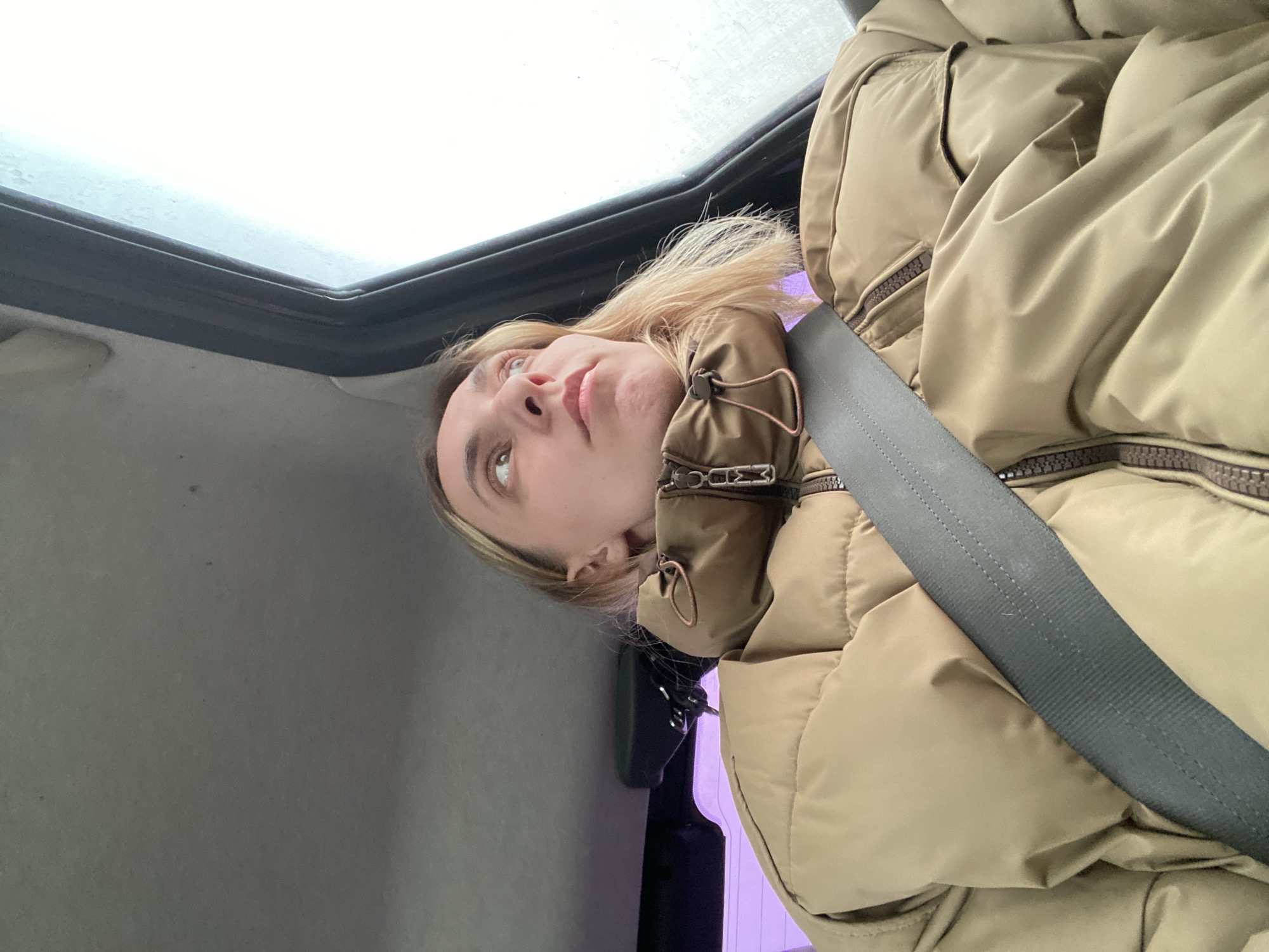 A woman is seated in a vehicle in Chaikovsky, Russia. She is wearing a tan puffer jacket and a seatbelt. A sunroof is visible above her, and part of the car's interior including a monitor can be seen in the background. The lighting suggests it is daytime.