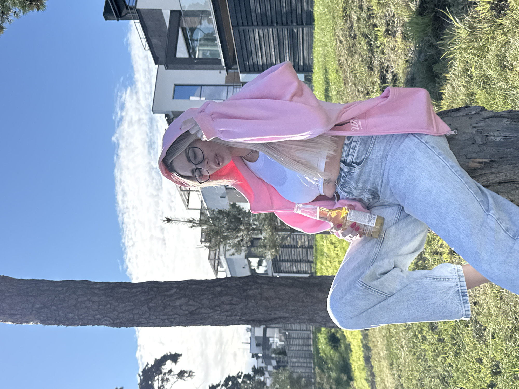 A blonde woman wearing glasses, a white crop top, light blue jeans, and a pink hoodie sits on a tree stump outdoors. She is holding a bottle of beer. The background shows a modern house with dark and white elements, a wooden fence, and green grass. The sky is blue with scattered clouds. The location appears to be a suburban area with residential buildings.
