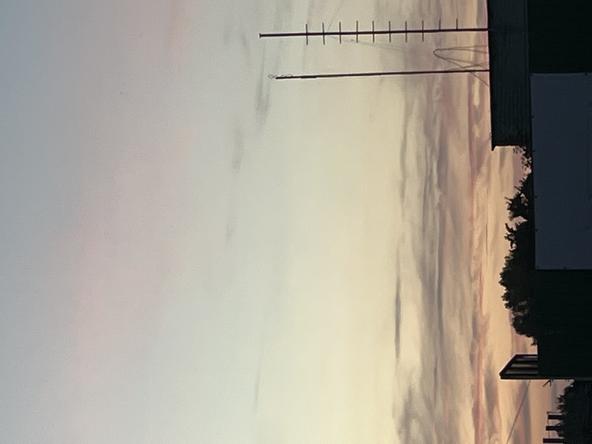 The image depicts a twilight sky over a cityscape. The sky exhibits soft hues of pastel pink, orange, and pale blue, indicative of sunset or sunrise. In the foreground, silhouetted structures of buildings and trees are visible, partially obscuring the view. Two tall radio towers, one with a ladder-like structure, stand prominently against the sky. The overall scene suggests an urban environment in Brest, Belarus, during the transitional light of dawn or dusk. No people or active interactions are discernable in the image.