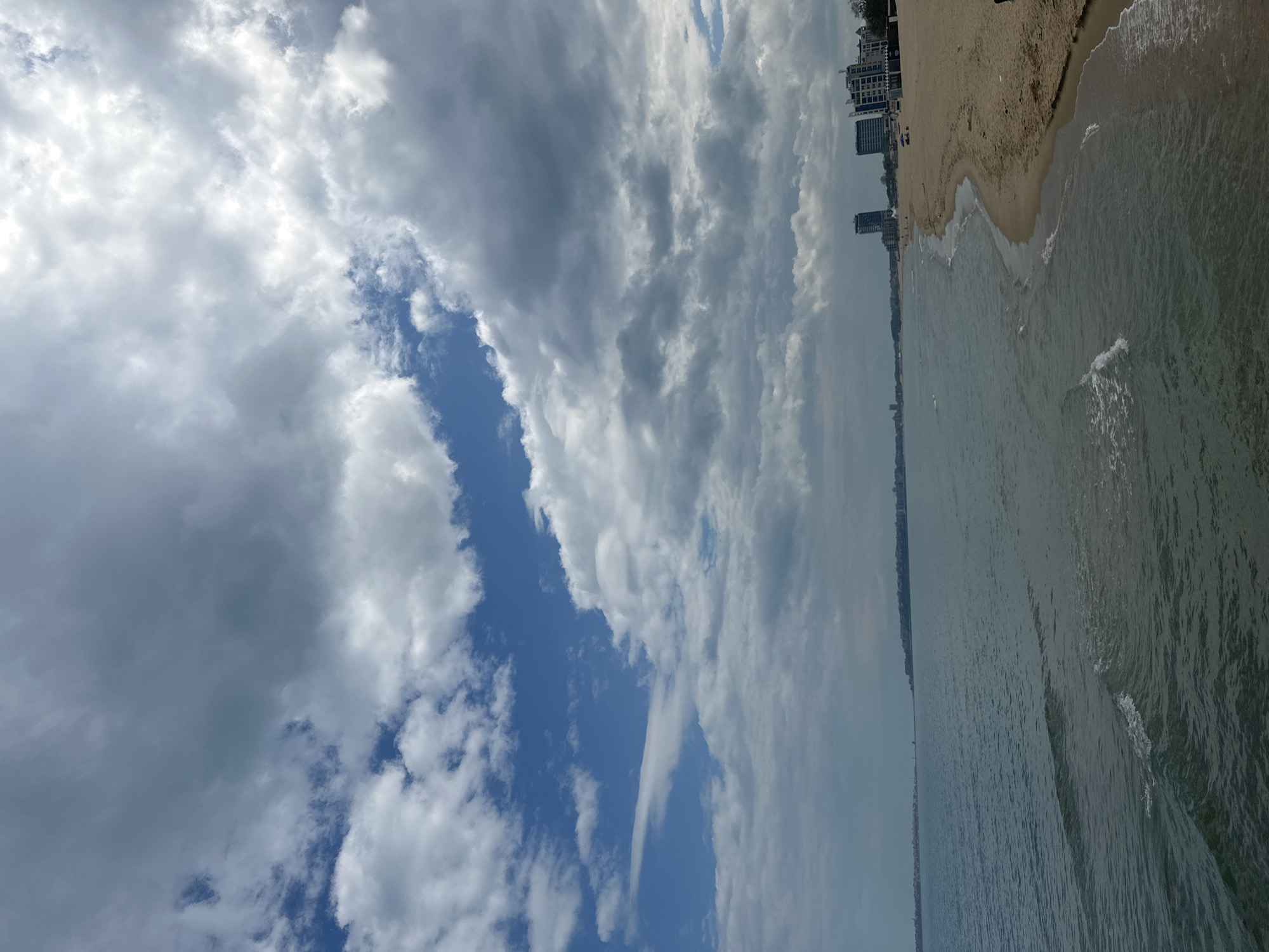 The image depicts a beach scene in Nesebar, Bulgaria. On the right side is a sandy beach with gentle waves lapping the shore. A row of buildings, likely hotels or apartments, can be seen along the beach. The sky is dominated by numerous clouds, varying in density from thick and dark to lighter, brighter patches, with some blue sky visible through the cloud cover. The image is taken from an elevated perspective, giving a wide view of the beach and sea.