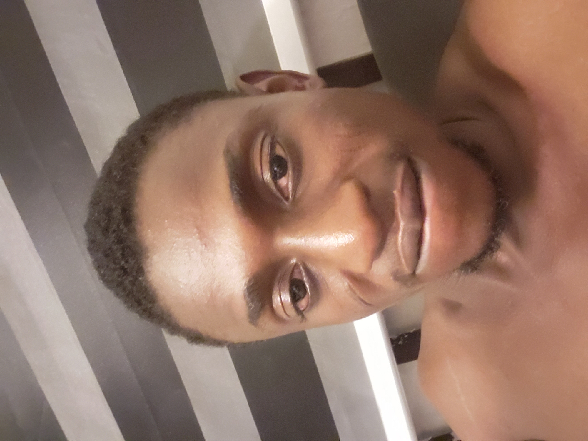 A close-up, slightly rotated selfie of a dark-skinned person with short, dark hair and a trimmed beard. The individual is smiling and appears to be well-lit. The background consists of black and gray vertical stripes, possibly wallpaper. The image provides no information to confirm a location in Adonkolo, Nigeria or any other specific location. The person is topless, revealing their shoulder. The scene appears to be indoors, possibly a bathroom or bedroom given the background.