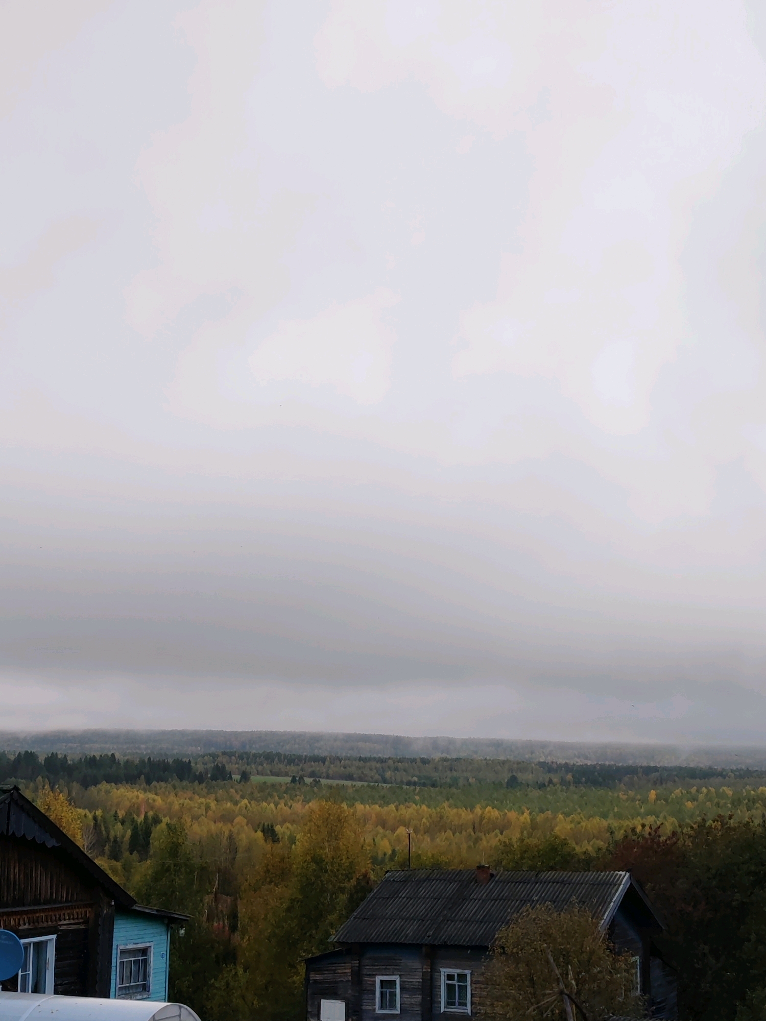 A high-angle, wide shot captures a hazy, overcast sky over a rural landscape. In the foreground, the roofs of two wooden houses are visible, one on the left with dark wood siding and a blue-painted section with a white window frame and a satellite dish. To the right, another house has a dark, corrugated roof and wooden siding with visible windows. Beyond the houses, a dense forest stretches to the horizon, its trees displaying a mix of green, yellow, and orange foliage, indicating autumn. The distant treeline is softened by a layer of fog or mist, and the sky is a uniform light gray. The overall impression is one of a quiet, rural setting in Russia, possibly in the village of Chukhlem, given the style of the houses and the natural landscape. There are no people or animals visible in the image.