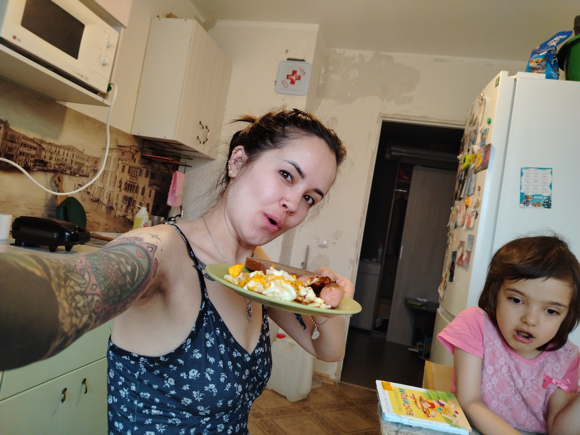 A medium shot depicts a woman taking a selfie in a kitchen while holding a plate of food, with a young girl seated at a table in the foreground.
The woman, with her hair pulled back, holds a plate of scrambled eggs, sausage, and a slice of bread. Her left arm, heavily tattooed, is extended towards the camera. She wears a floral patterned tank top and a silver necklace. Multiple piercings are visible on her left ear.
A young girl with dark hair sits at a table in the lower right corner. She is wearing a pink lace top.
The kitchen includes light-colored cabinets, a microwave, a refrigerator covered in magnets, and a first aid kit on the wall. A partial view of a room is visible through an open doorway behind the refrigerator. A book rests on the table near the girl.
The location is likely in a residential setting in the city of Noviy, Russia, based on the book's title in Russian.