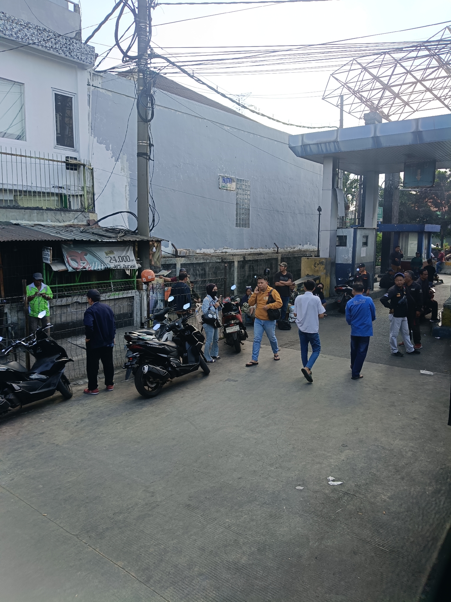 The image depicts a street scene in Kedungrejo, Indonesia, near a gas station. Several people, mostly men, are visible. Some are standing and talking, while others appear to be waiting or walking. There are several motorcycles parked on the side of the road. A small building with a corrugated metal roof is also present, with signage indicating it may be a shop or kiosk. A gas station canopy with visible pumps is in the background. The overall environment suggests a bustling urban or semi-urban area.