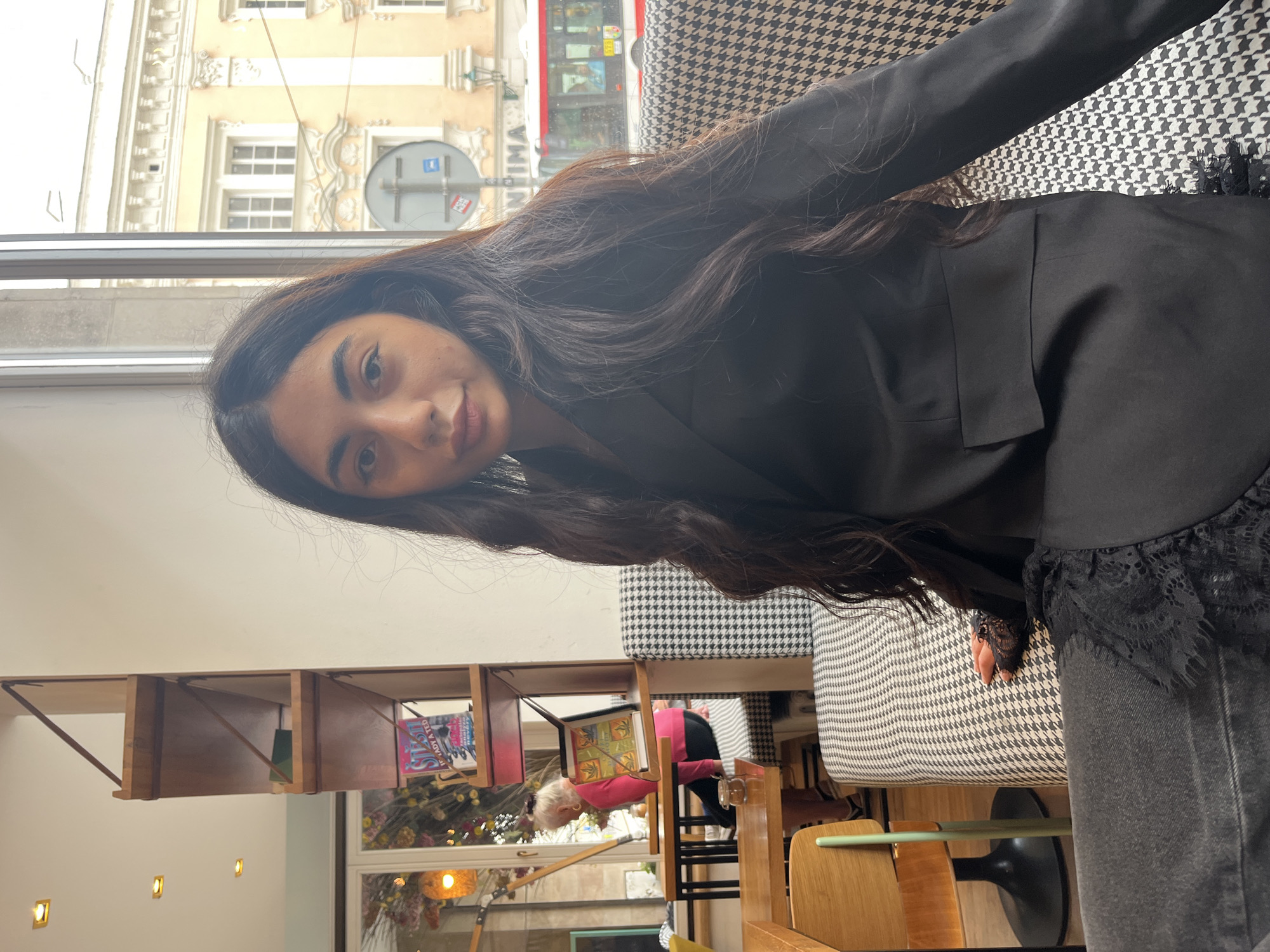 A young woman with long, dark hair is seated at a table in a cafe. She is wearing a black blazer over a black top with lace trim, and dark denim jeans. She is looking towards the camera. In the background, a woman with blonde hair wearing a pink top is seated at another table, and there are shelves with books and decorative items on the wall. Through a large window, a bus and buildings in the city of Pilsen, Czechia, are visible. The cafe has black and white patterned seating.