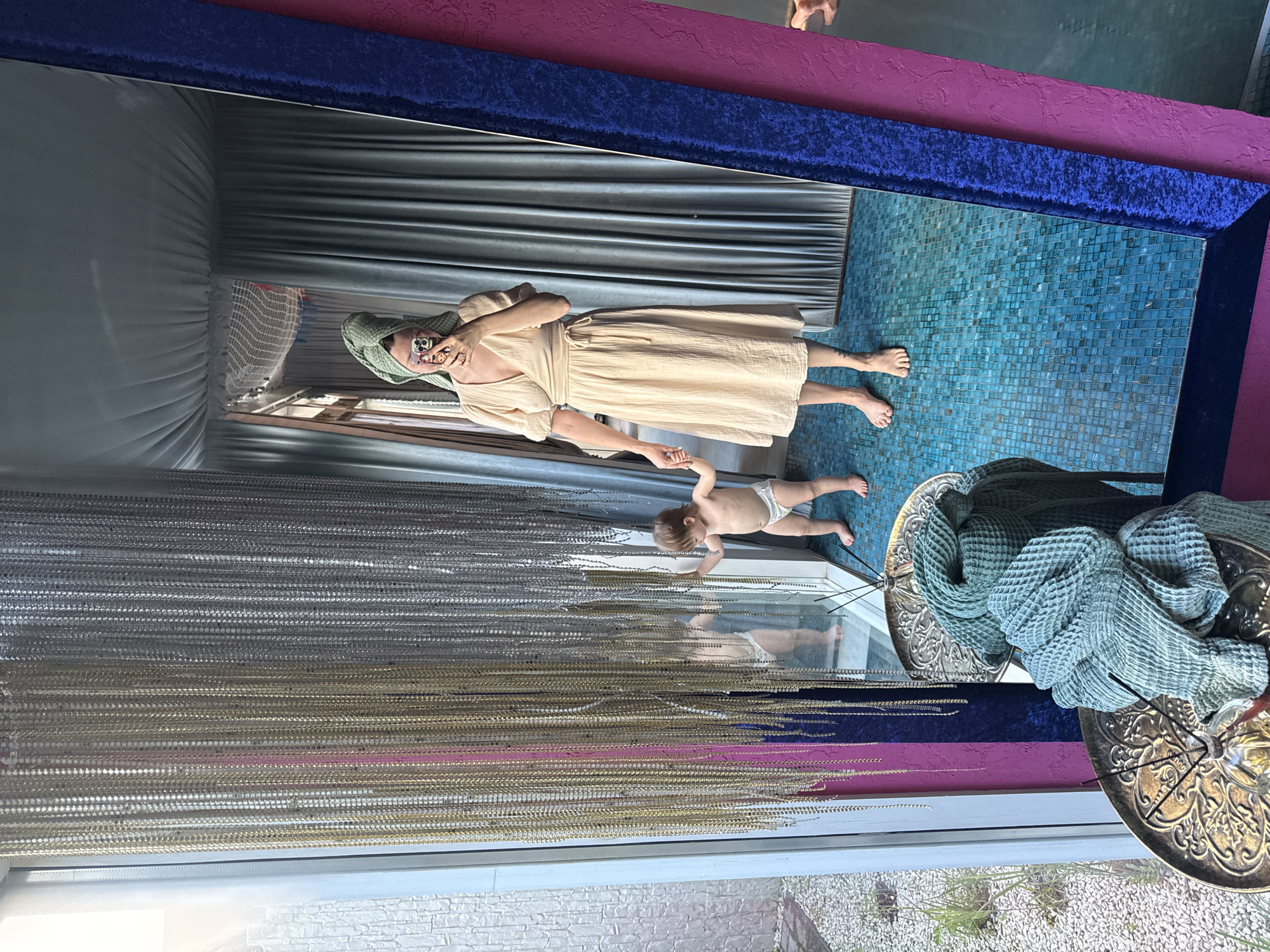 The image shows a reflection in a mirror with a blue and purple frame. A woman wearing a light-colored dress and a head wrap is standing on a blue tiled surface and holding a smartphone to take a picture. A child in a diaper is reaching towards the woman. The reflection shows a gray curtain behind the woman and a draped netting structure. The foreground of the image shows a window with beaded strands and the exterior featuring small pebbles and vegetation. The location of Чёрное, Russia is not discernible from the visual information.