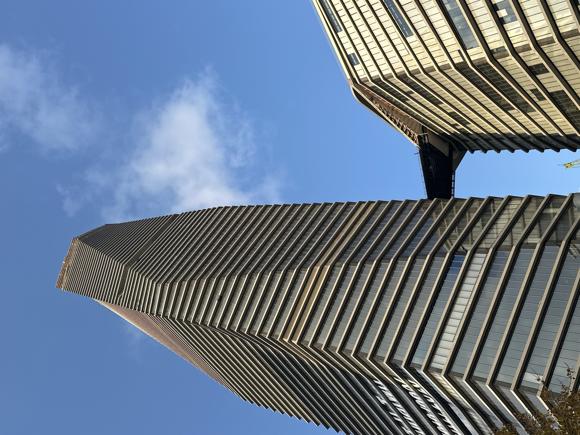 The image captures a dynamic, low-angle perspective of two striking modern skyscrapers against a brilliant blue sky dotted with sparse white clouds. The entire scene is tilted, enhancing the sense of height and architectural grandeur.
The prominent building on the left exhibits a unique, undulating facade composed of numerous stepped and angled panels, creating a repetitive, ribbed pattern that gives the structure a fluid, almost organic appearance as it ascends. Its surfaces appear to be a mix of glass and light-toned materials, reflecting the daylight.
Partially visible on the upper right is a second, equally tall building, characterized by a more conventional, grid-like facade with numerous windows. A dark, multi-level skybridge or connecting structure spans between the upper sections of the two buildings, visually linking them. A faint yellow construction crane is visible near the upper right edge, suggesting ongoing development.
The bright daytime sun illuminates the buildings, casting distinct shadows that emphasize the depth and architectural features. A small cluster of dark tree foliage can be seen in the bottom right corner of the frame. The clear sky and strong lighting indicate fair weather. This impressive architectural landscape is located in Tel-Aviv, Israel. No visible text is present in the image.
