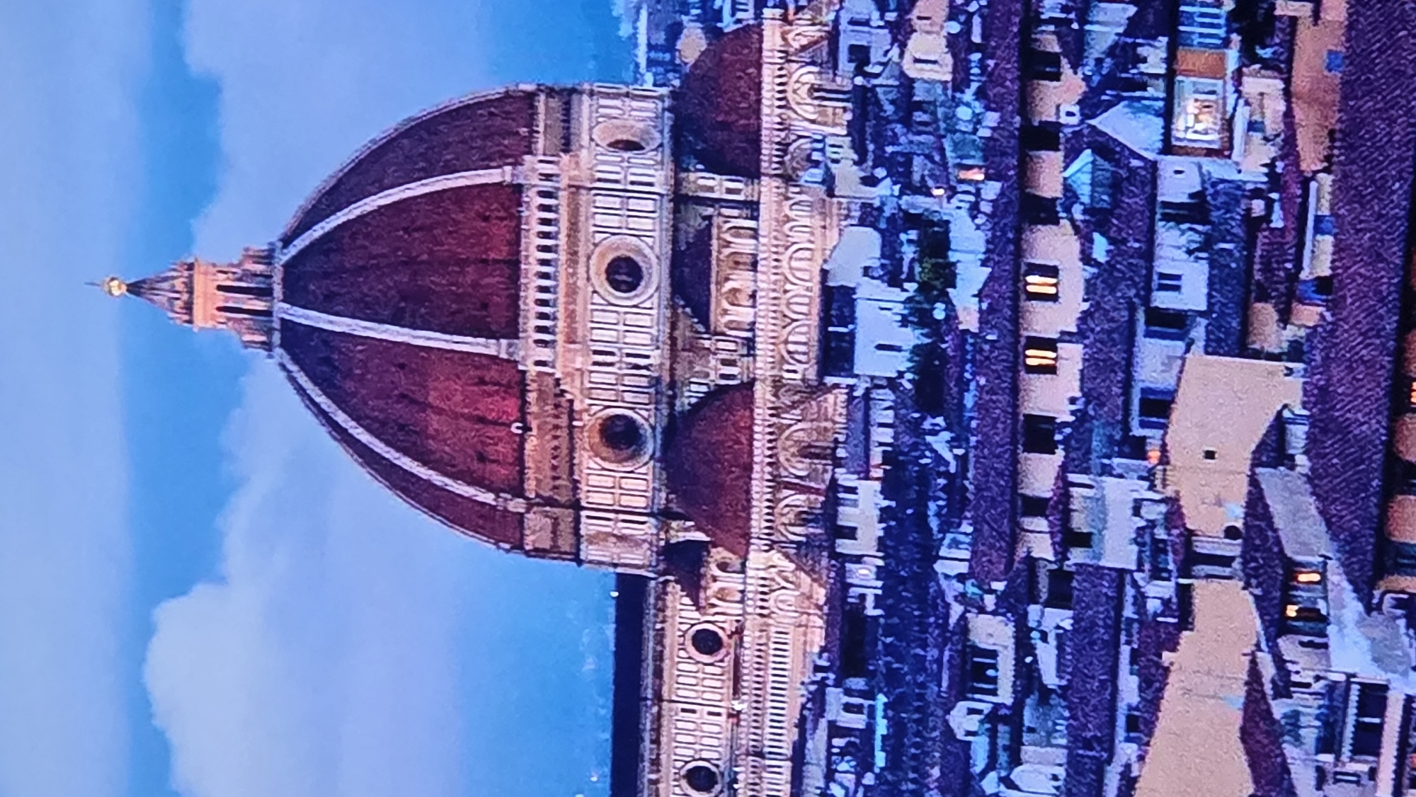The image displays a close-up view of the Florence Cathedral dome and surrounding cityscape. The dome is a dominant feature, with its reddish-brown tiles and white vertical accents, topped by a smaller lantern. Below the dome, the cathedral's intricate facade is visible, featuring arches and circular windows. The foreground is filled with a dense collection of buildings with terracotta roofs, characteristic of a European city. Some windows of these buildings are illuminated, suggesting it is evening or night. The sky above is a deep blue with soft, cloudy textures. The scene is a picturesque panorama of a historic Italian architectural marvel and its urban environment.