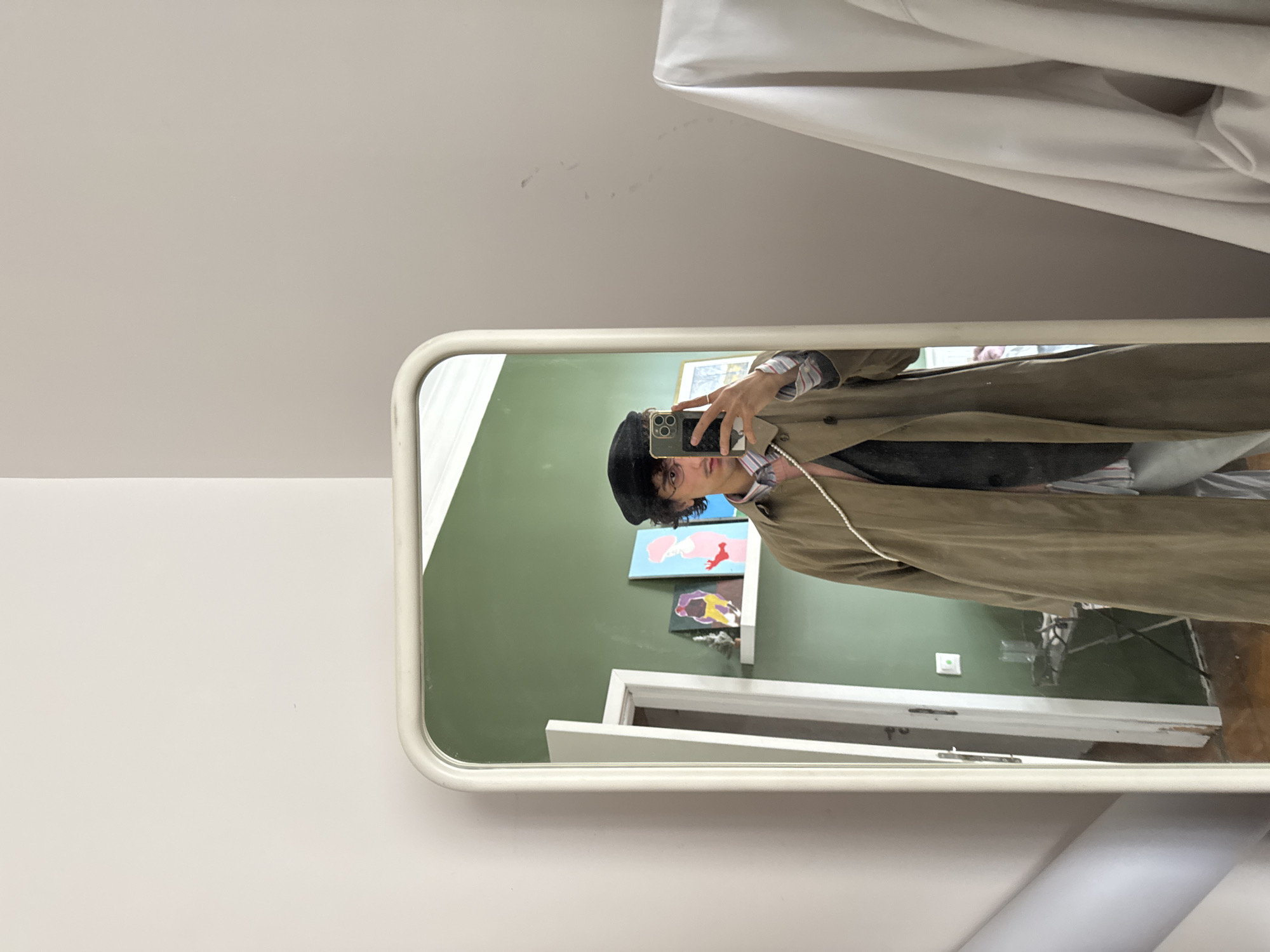 A person takes a selfie in a mirror. The person is wearing a black beret, glasses, a collared shirt, a vest, and a tan coat. Behind the person, inside the room, is a green wall with two pieces of art on it. A door frame is visible. A microphone on a stand is also visible. The location is not identifiable as Moscow, Russia.
