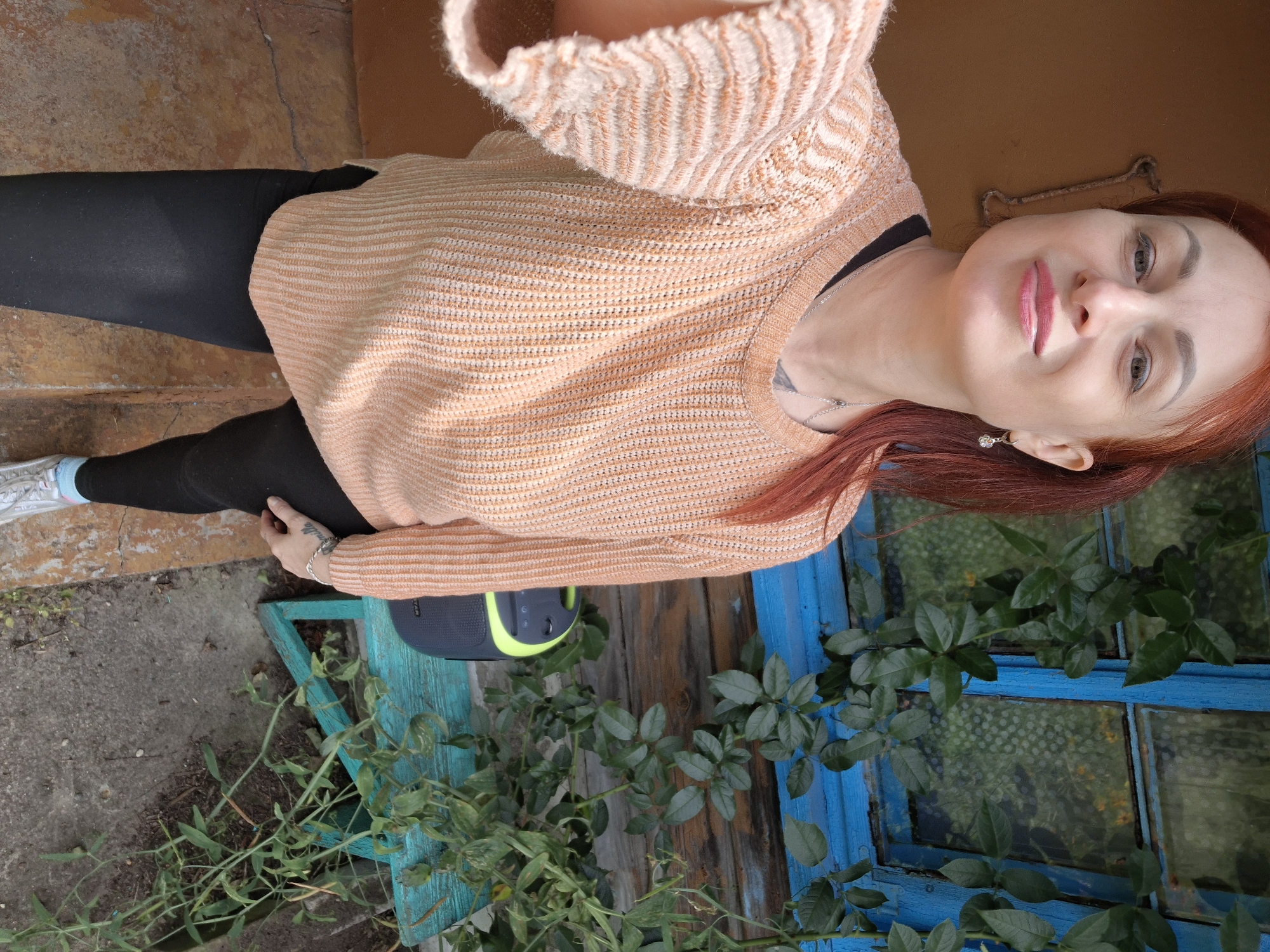 A woman with red hair stands on a concrete and dirt path in front of a rustic wooden building with a blue-framed window. She is wearing a peach-colored knitted sweater, black leggings, and white sneakers with blue accents. A small silver necklace with a pendant is visible around her neck. To her right, a small blue bench holds a portable speaker with a black and green design. Green vines with leaves grow around the window and down the side of the building. The overall scene suggests a rural or semi-rural setting, possibly a home or outbuilding.