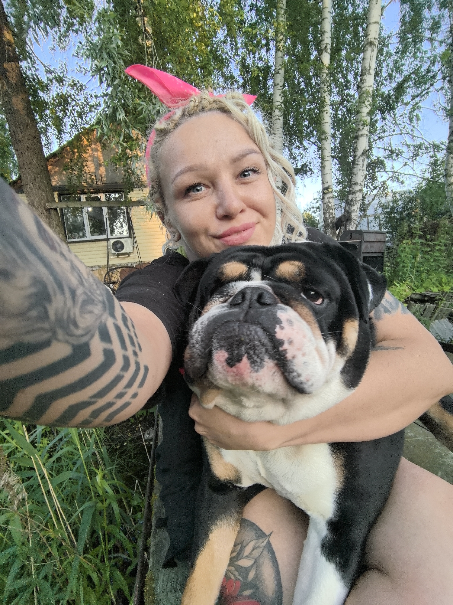 A woman with blonde curly hair and a pink headband is holding a black, white, and tan bulldog. She is wearing a black t-shirt and has extensive tattoos on her left arm. The bulldog is looking towards the camera. They are outdoors, with trees and a small yellow building in the background. The setting appears to be a rural or suburban area. The image is a selfie, taken from a slightly low angle. The woman's right arm is wrapped around the bulldog, and her left arm is also visible, with the tattoos being a prominent feature. The bulldog's face is close to the woman's face. The background shows green foliage and the building has windows and an air conditioning unit. The sky is visible above the trees, and it appears to be daytime.
