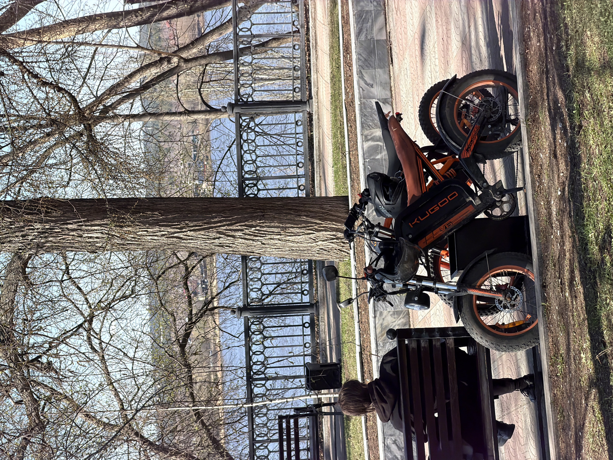A vertical, full outdoor shot captures a person seated on a park bench, adjacent to a KuGo electric bike, likely in Perm, Russia. The bike is black and orange, with a headlight and mirrors. A helmet is secured to the handlebars. The park bench is wooden with a black metal frame. The person sitting on the bench is wearing dark clothing.
The background features a black metal fence through which a body of water can be seen. Trees, with bare branches suggesting it may be early spring, frame the view. The ground is paved with light colored stones on one side and grass on the other.