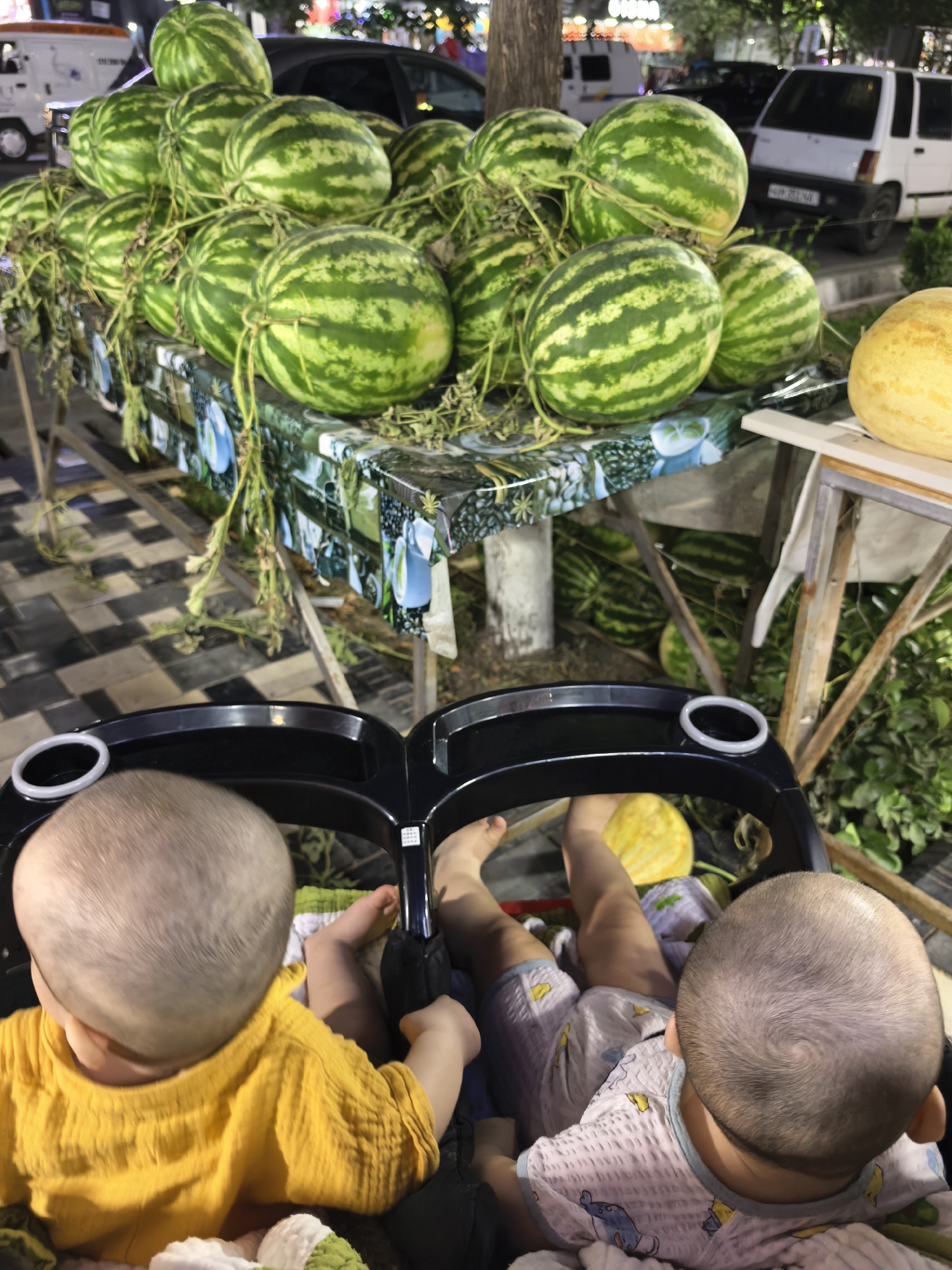 A street-side market stall in Fergana City, Uzbekistan, displays a large quantity of watermelons and several melons. The watermelons are stacked on a table covered with a patterned cloth. Some watermelons have vines attached, suggesting they are freshly harvested. Behind the stall, a white car and a dark-colored car are visible, along with some greenery. In the foreground, two infants are seated in a stroller, facing away from the camera. The infant on the left wears a yellow shirt, and the infant on the right wears a patterned outfit. The stroller has black framing and cup holders. The ground appears to be paved with a checkered pattern. The lighting suggests it is evening.