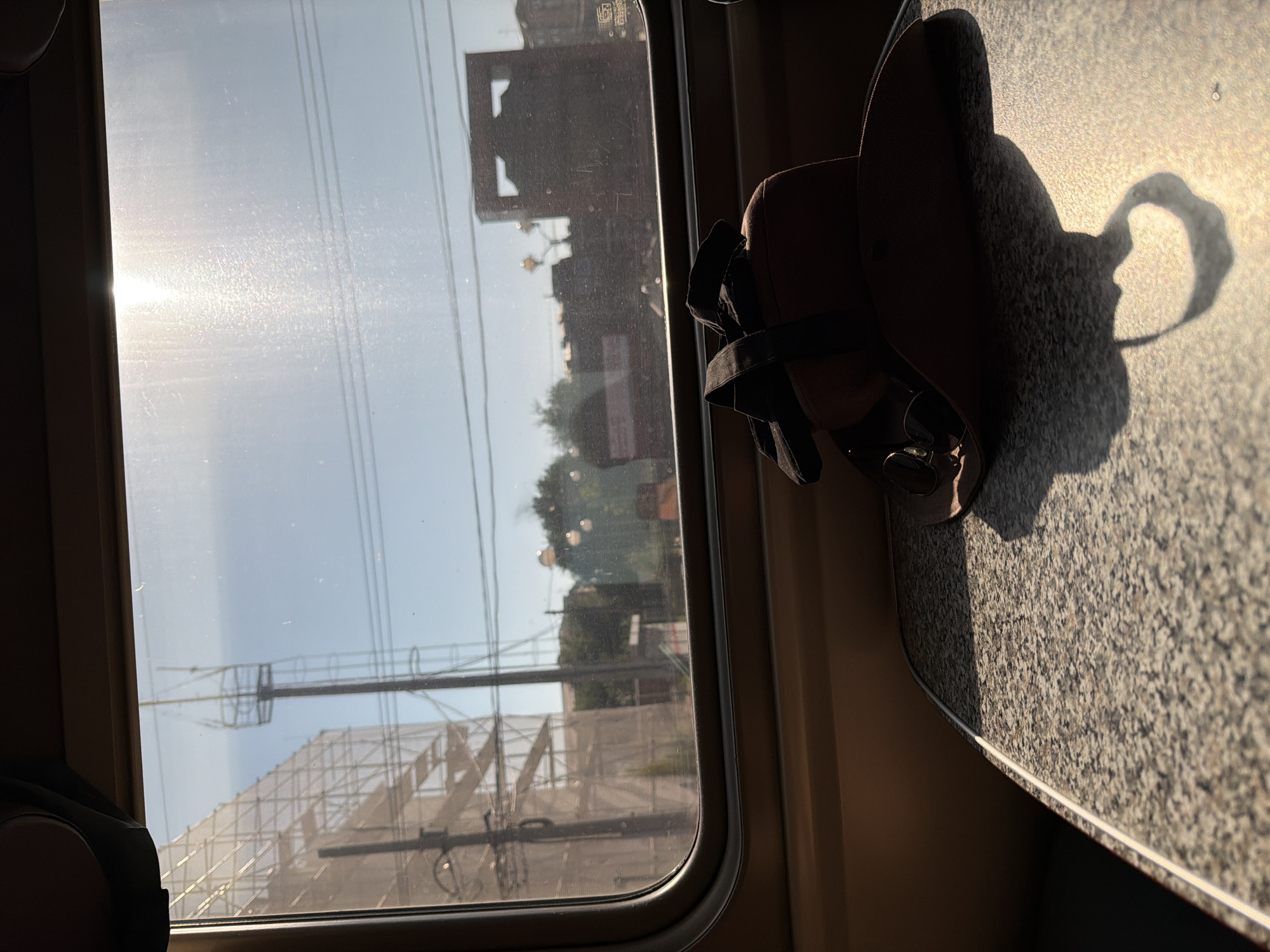 The image depicts a view from a train window, looking out onto a cityscape. In the foreground, on a speckled tabletop, rests a brown hat with black straps and a pair of sunglasses tucked inside. The hat casts a distinct shadow on the table. Outside the window, the bright sun creates a lens flare, obscuring some details. Visible in the background are buildings, trees, utility poles, and scaffolding. The environment suggests a sunny day, possibly in an urban or semi-urban setting. The location is identified as Petrov Val, Russia. There are no people visible in the image.