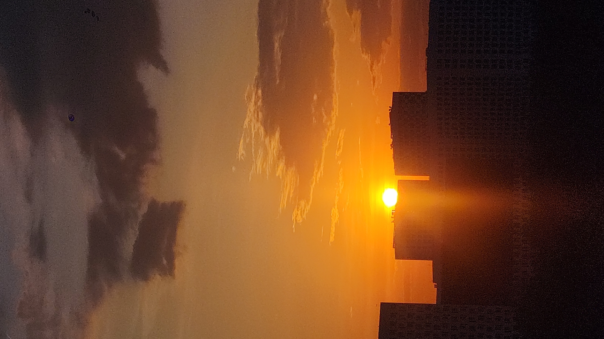 The image shows a sunset over Moscow, Russia. The sky is filled with clouds that are colored orange and yellow by the setting sun. The sun itself is visible as a bright yellow orb. The silhouettes of buildings can be seen in the foreground.