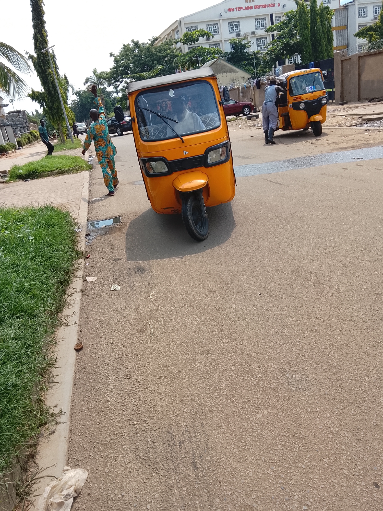 In the city of Abuja, Nigeria, a bright orange auto-rickshaw is parked on a paved road. Another similar auto-rickshaw is visible further down the road. A man in a patterned tunic and trousers stands on the sidewalk, raising his right arm. Beside him, a person in a grey shirt and trousers is interacting with the parked auto-rickshaw. A black car is parked across the street, and a building with a sign reading "WHITIPLANS BRITISH SCHOOL" is visible in the background. Green grass lines the edge of the sidewalk. The scene suggests an urban street environment with moderate activity.
