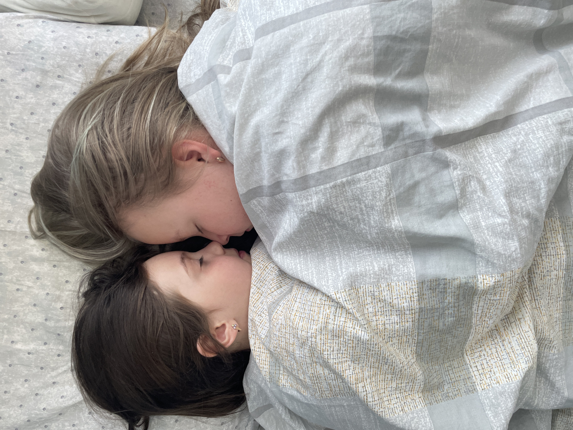 Two individuals are lying head-to-head on a bed covered with bedding. One individual has dark hair, and the other has blonde hair. They appear to be kissing or in close contact. A patterned blanket covers part of their bodies. The location is not determinable from the image and the request to include Izhevsk, Russia, is not supported by the image.